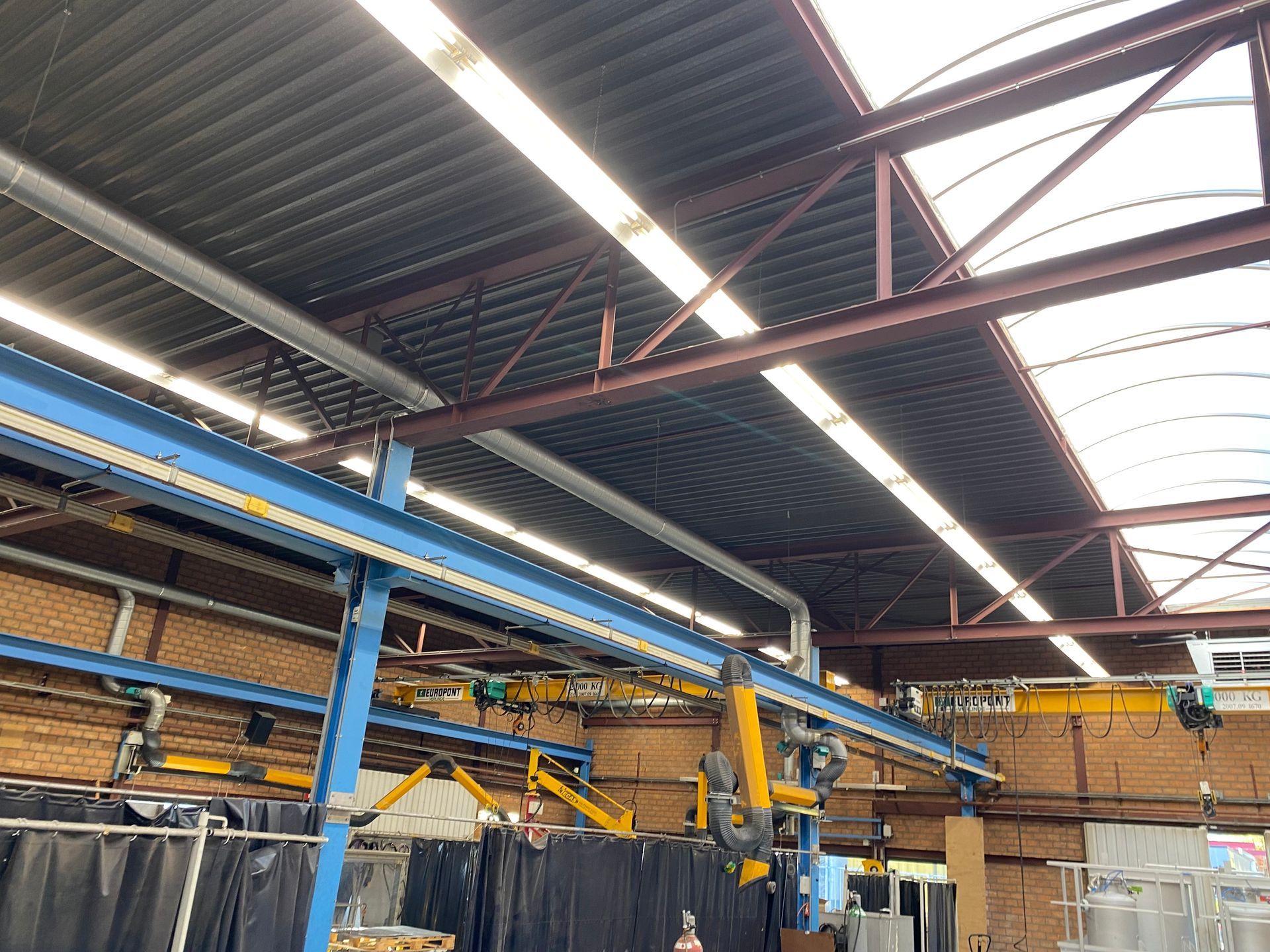 Interieur van een industriële werkplaats met hoge plafonds, zichtbare spanten, lichte dakramen en gele brugkranen.