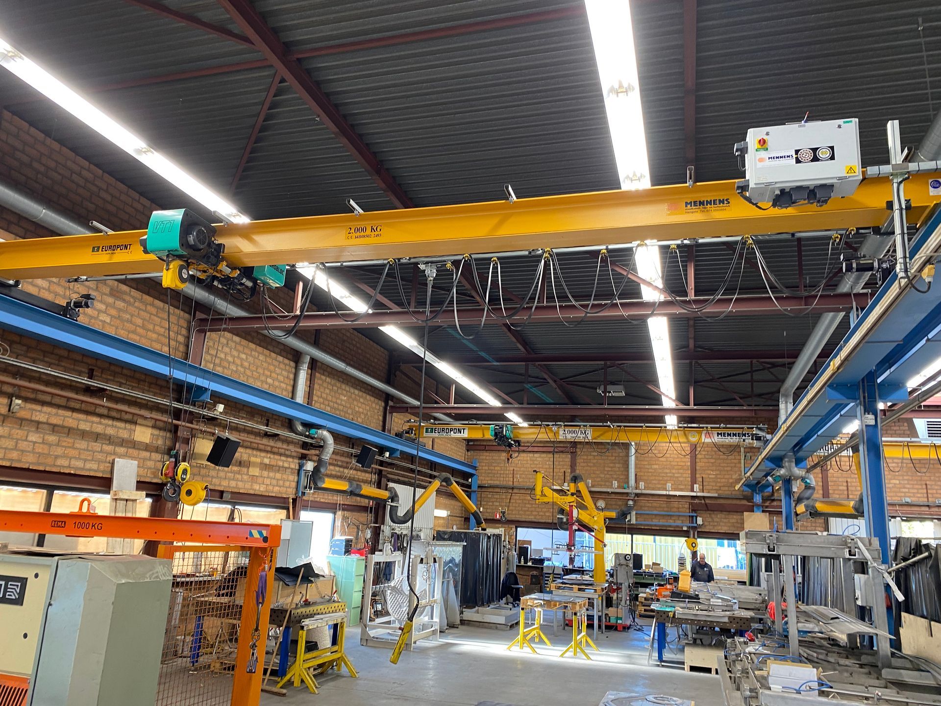 Een industriële werkplaats met gele bovenloopkranen, gemonteerd op blauwe stalen steunpilaren die de hele constructie overspannen.