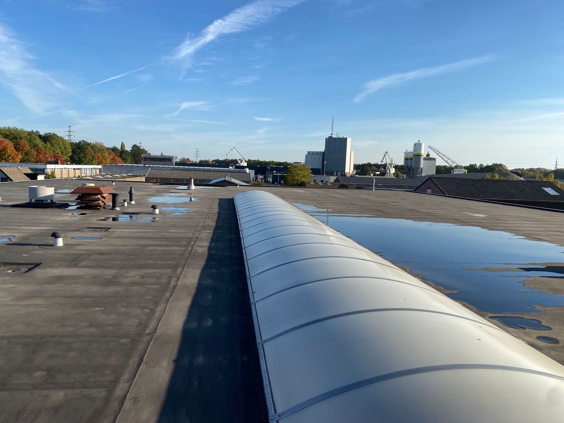 Plat industrieel dak met een lang, gebogen dakraam en plassen stilstaand water onder een helderblauwe hemel.