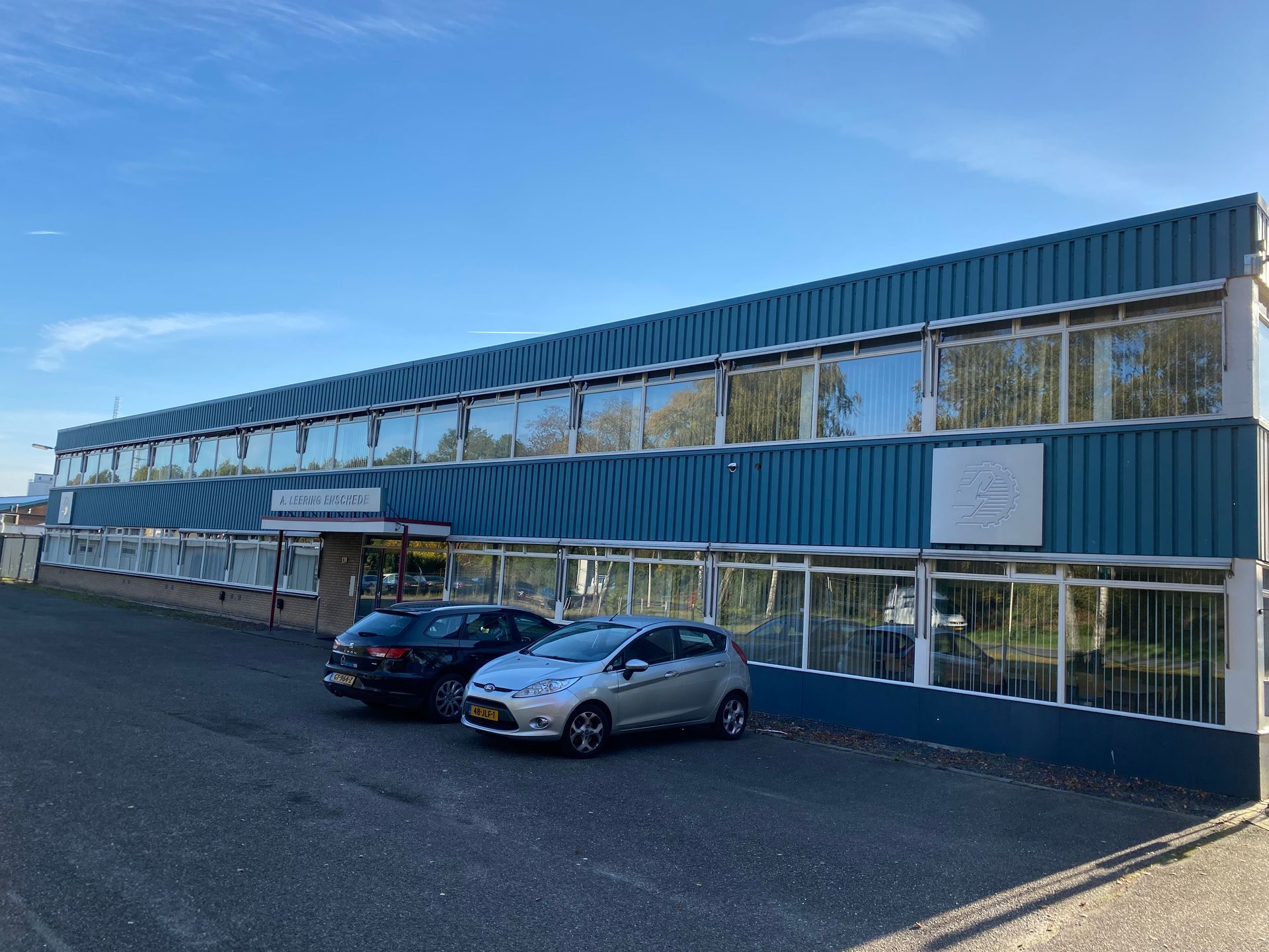 Een blauw industrieel gebouw van twee verdiepingen met grote ramen en een parkeerplaats ervoor met twee auto's, onder een helderblauwe hemel.