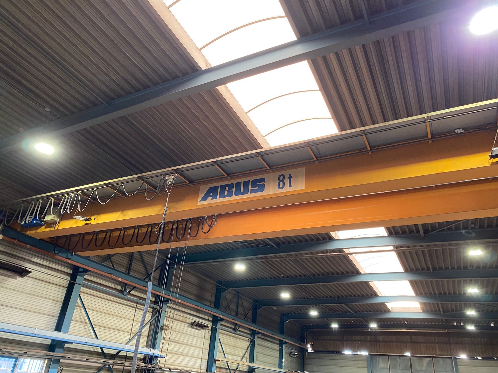 Een oranje ABUS 8-tons bovenloopkraan, gemonteerd op plafondbalken in een industriële loods met dakramen.
