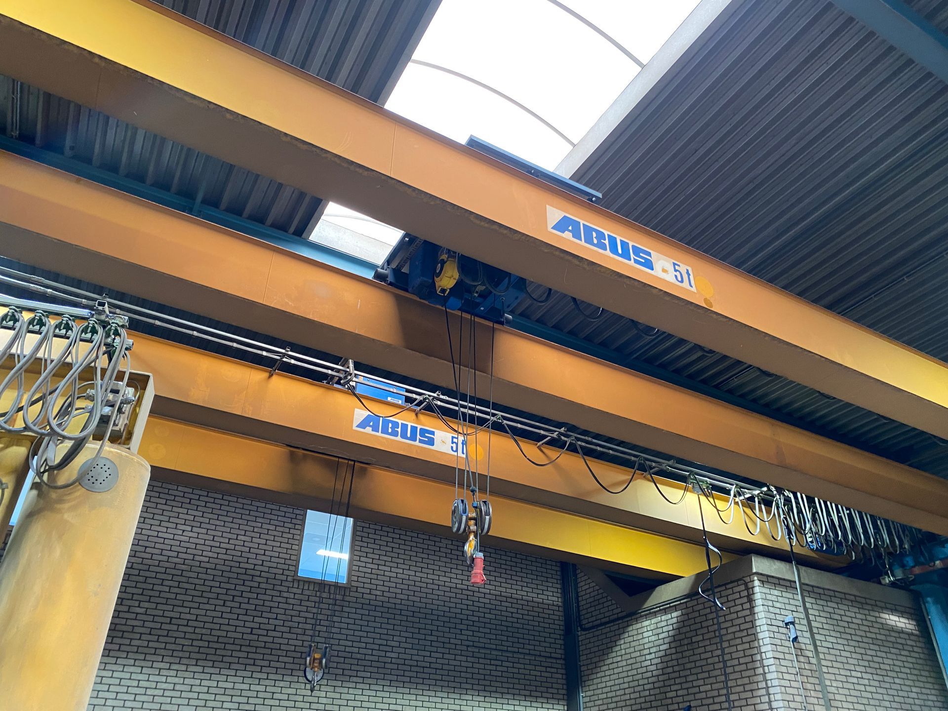 Gele bovenloopkraan met een hijseenheid die aan metalen balken is opgehangen in een industriële loods.