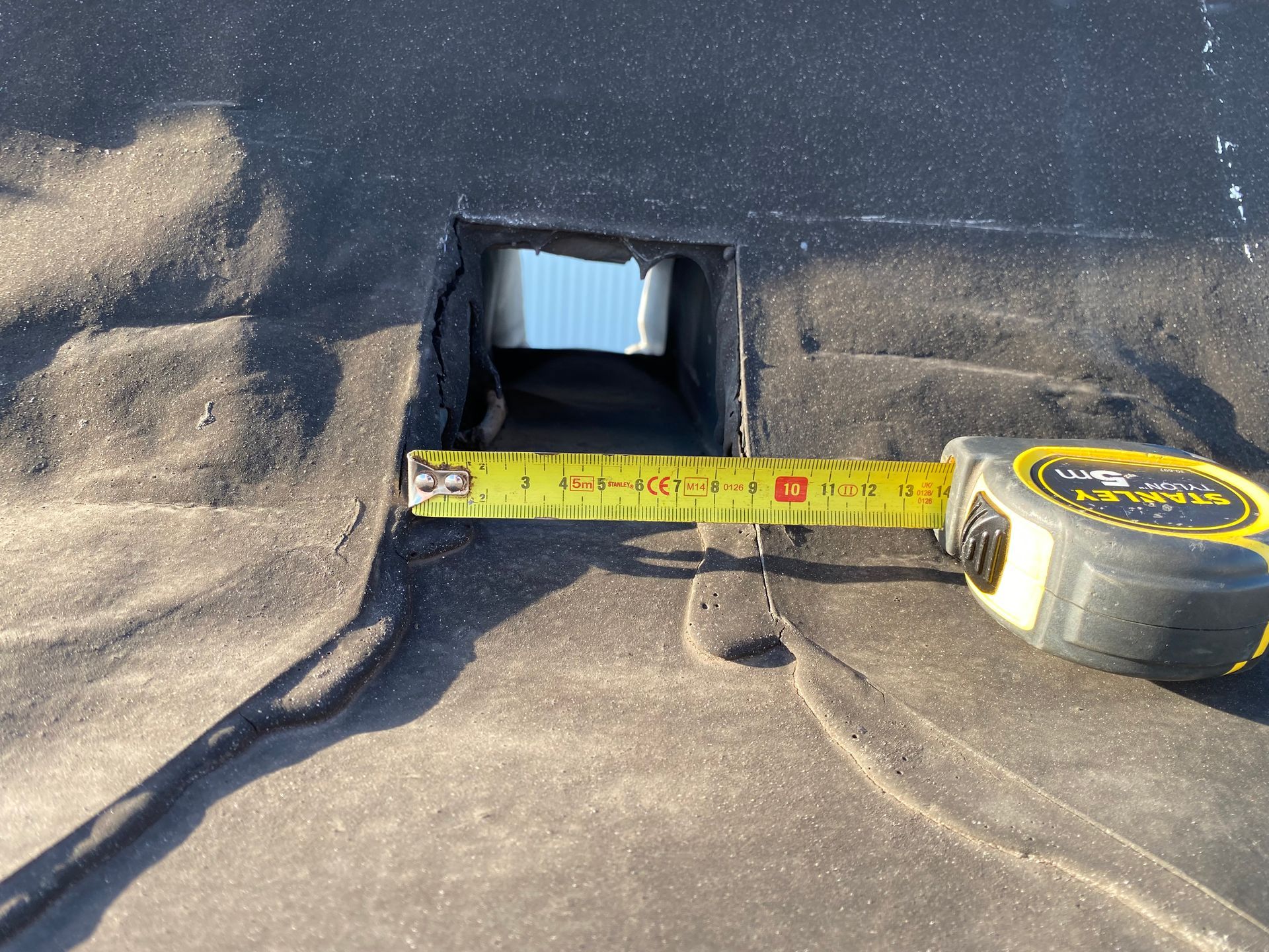 Een meetlint laat een rechthoekig gat zien dat in een zwart plat dak is gemaakt, waardoor de glanzende metalen luchtkanalen eronder zichtbaar worden.