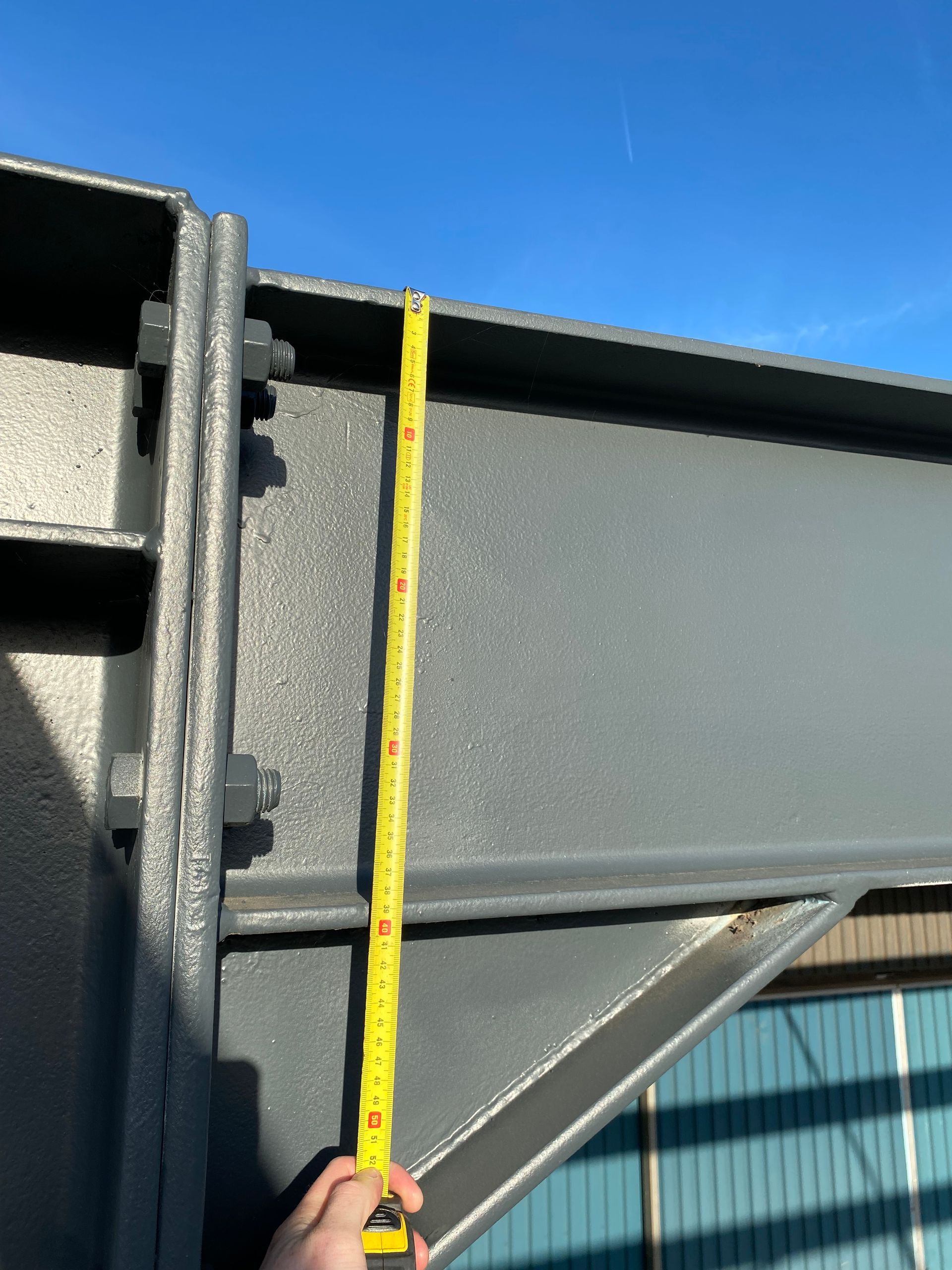 Een hand houdt een geel meetlint tegen een grijze stalen balk en de bijbehorende steunbeugel, buiten onder een blauwe hemel.