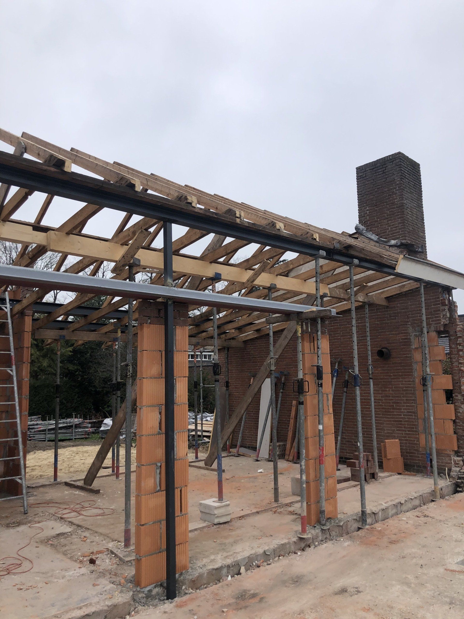 Bouwplaats met een bakstenen constructie en een zichtbaar houten dakframe, ondersteund door metalen balken en steigers.