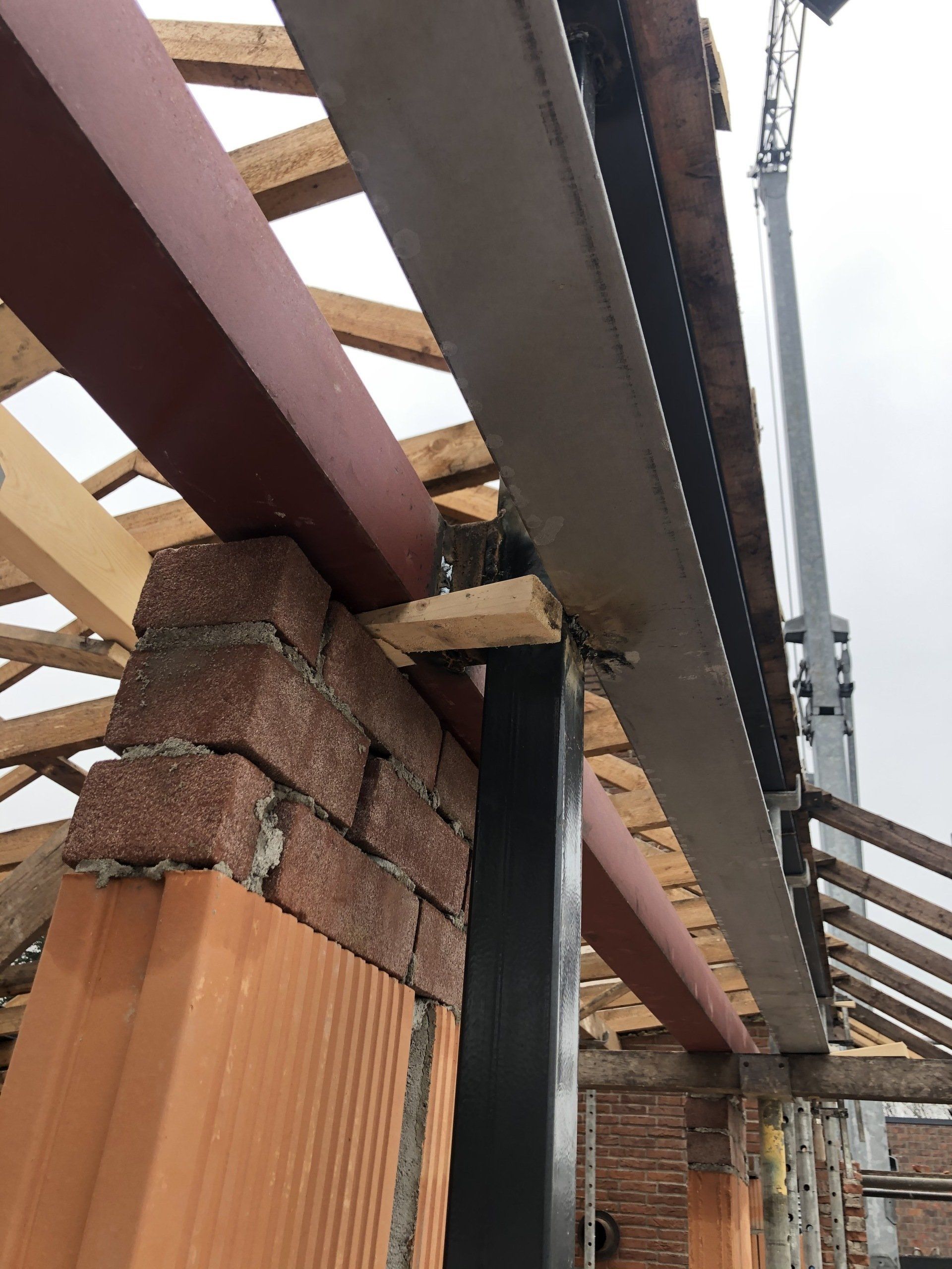 Bouwtekening van een stalen balk, ondersteund door een bakstenen kolom en een houten frame, met een kraan zichtbaar op de achtergrond.