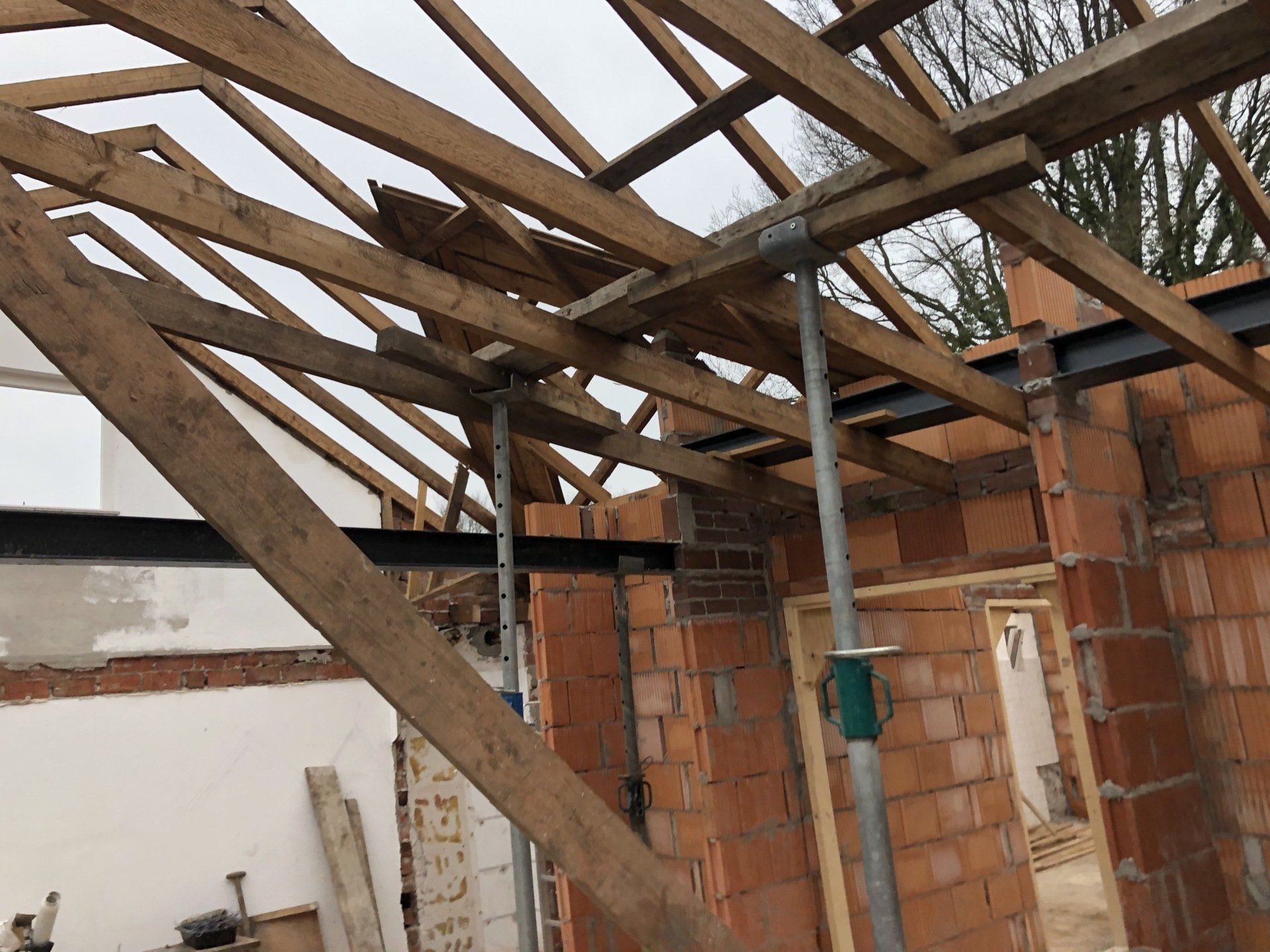 Interieur van een bouwplaats met zichtbare houten dakspanten, ondersteund door tijdelijke metalen palen boven een stalen balk.