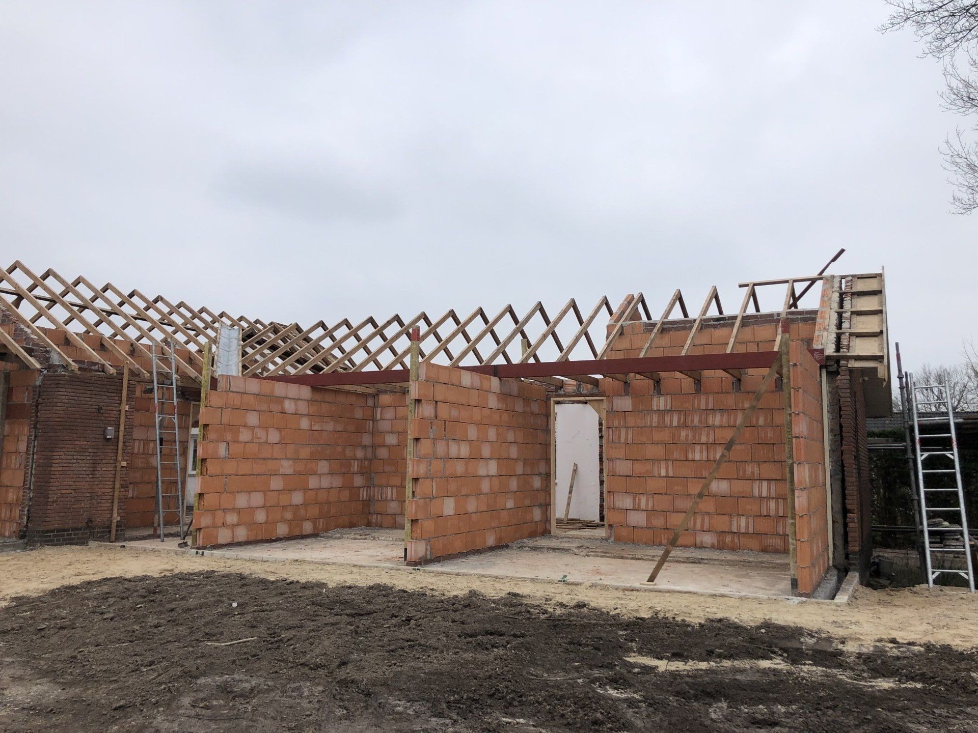 Op een bouwplaats is een eenlaags bakstenen gebouw te zien met een zichtbaar houten dakspant onder een bewolkte hemel.