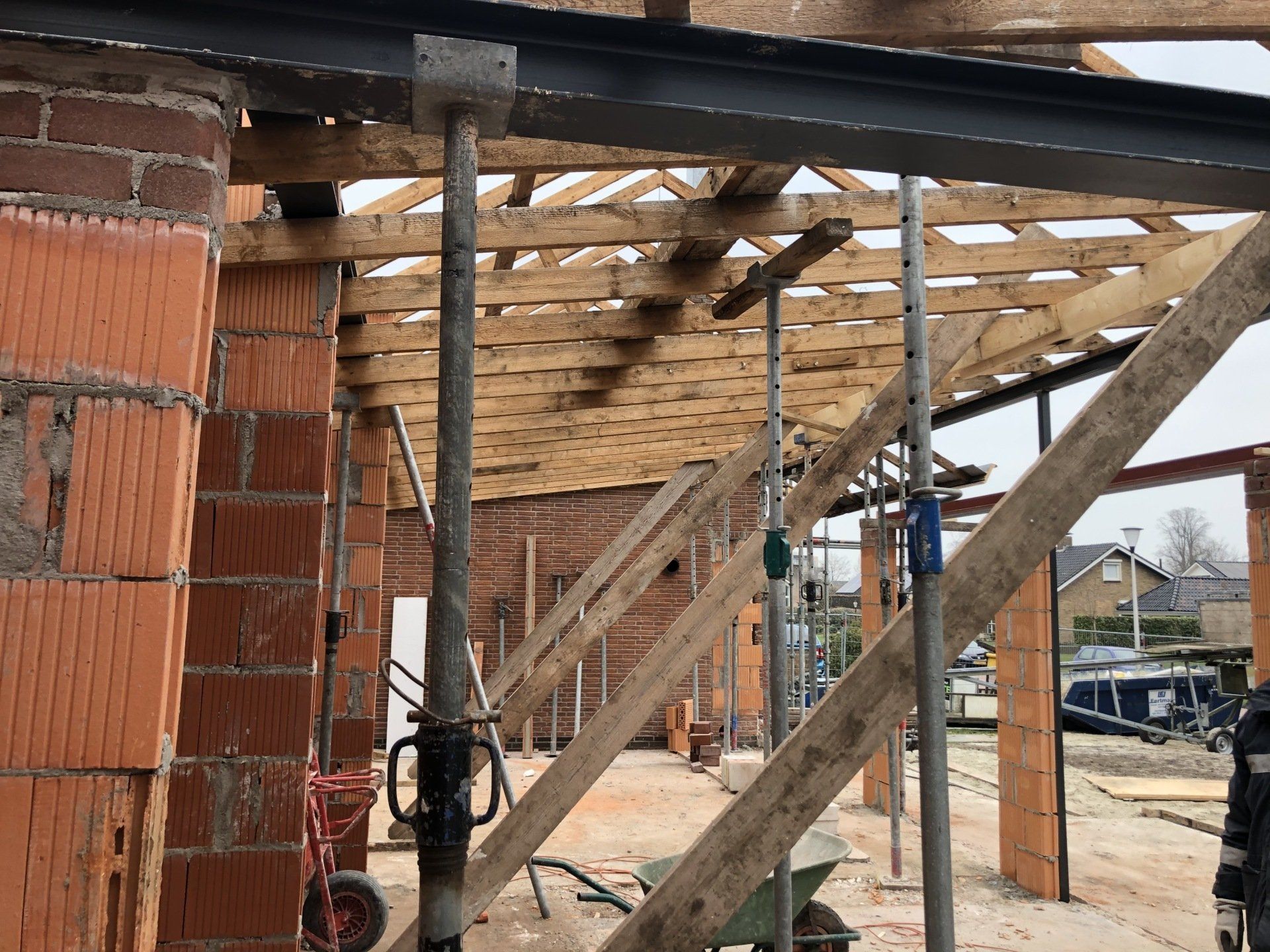 Op een bouwplaats is een stalen balk te zien die wordt ondersteund door verstelbare metalen stutten onder een houten dakconstructie.