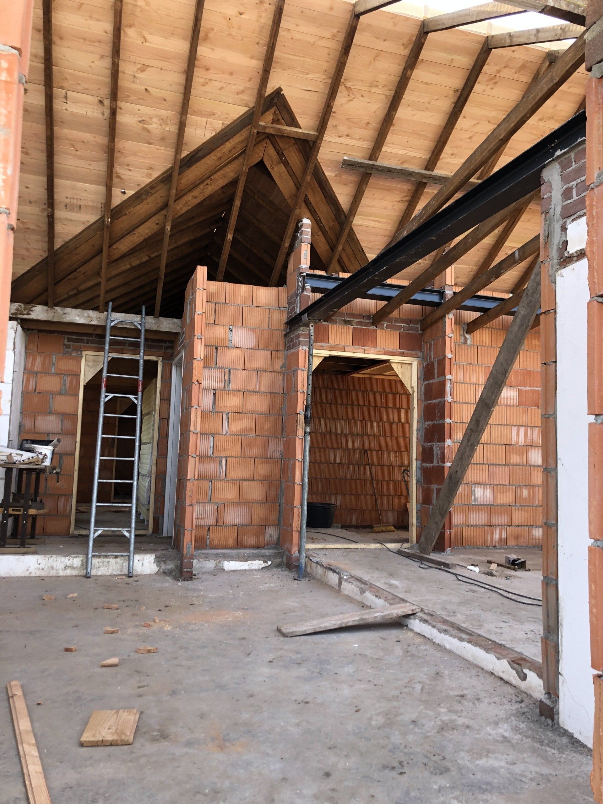 Een bouwplaats binnenin met rode bakstenen muren, een open houten dakconstructie en een metalen ladder in een deuropening.