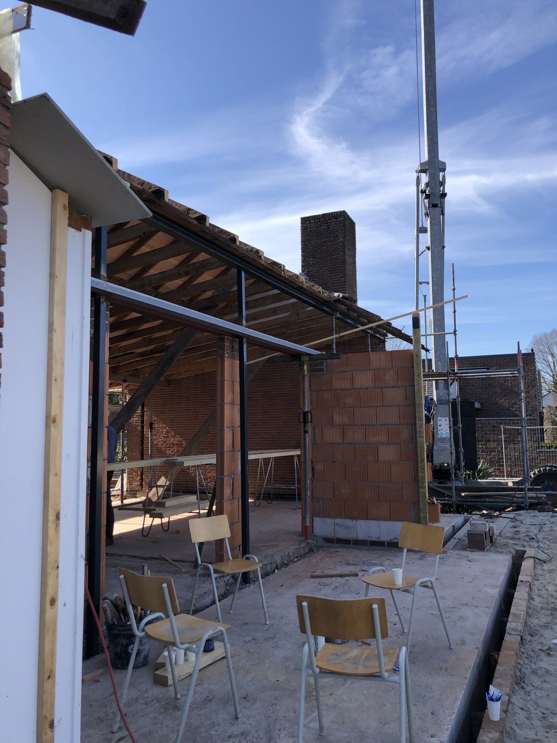 Een bouwplaats met vier houten stoelen voor een open, half gerenoveerd gebouw onder een blauwe hemel.