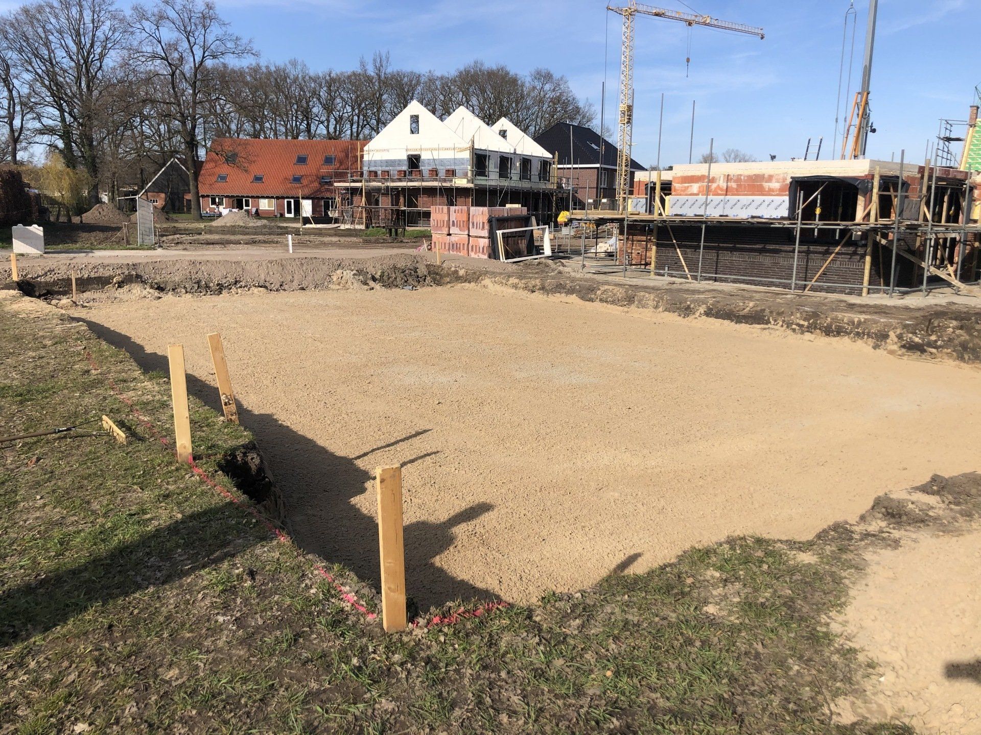 Een vlakke, met zand gevulde fundering van een bouwterrein, afgebakend met houten palen, met onafgewerkte huizen op de achtergrond.