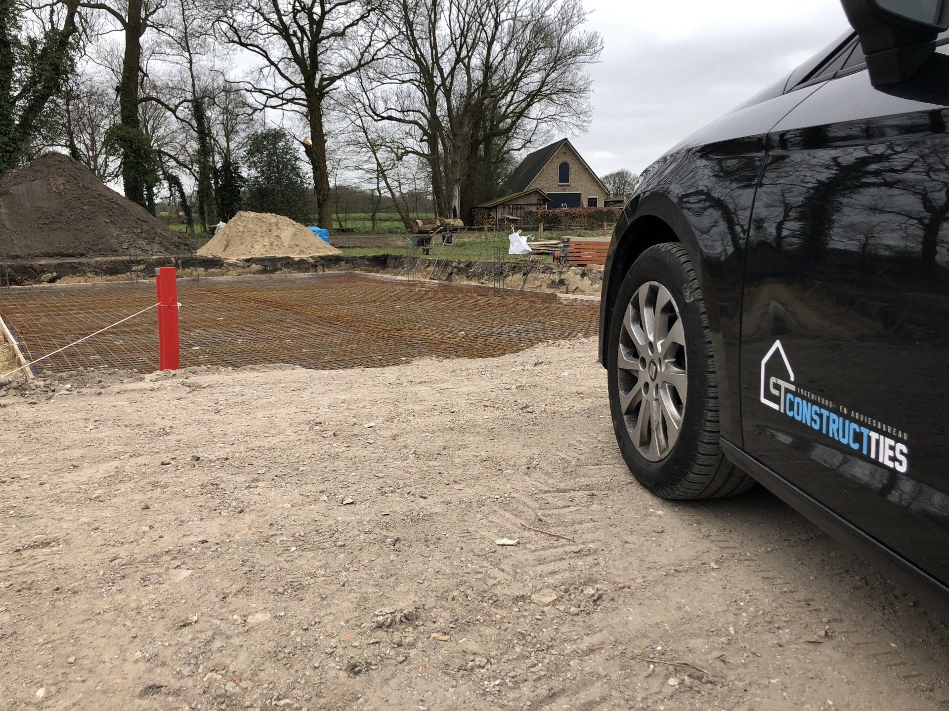 Een zwarte bedrijfsauto geparkeerd naast een bouwterrein met hopen grond en een huis op de achtergrond.