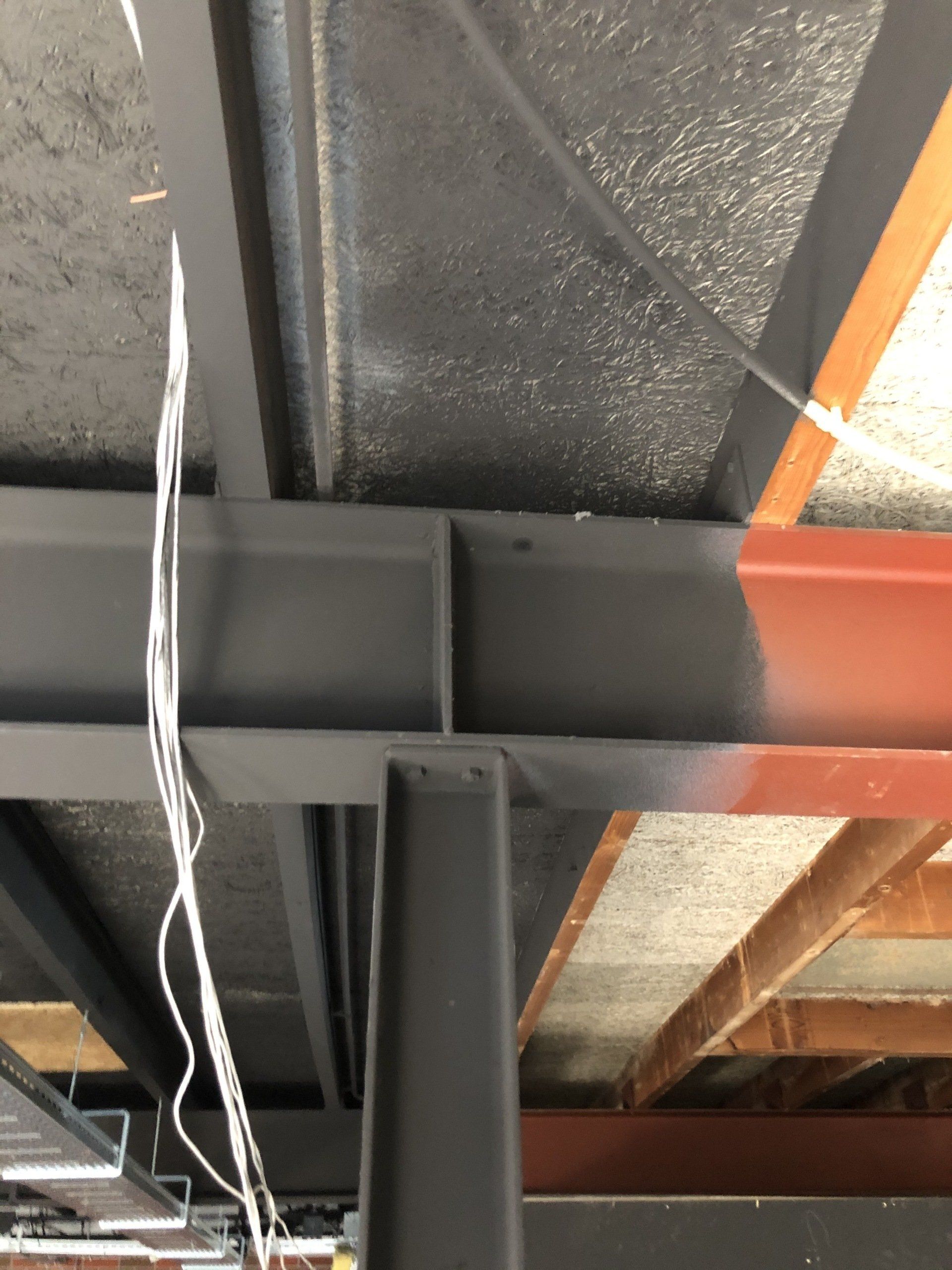 Een zwarte stalen I-balk kruist een andere, die gedeeltelijk in bruine grondverf is geschilderd, met daarboven de zichtbare plafondconstructie.