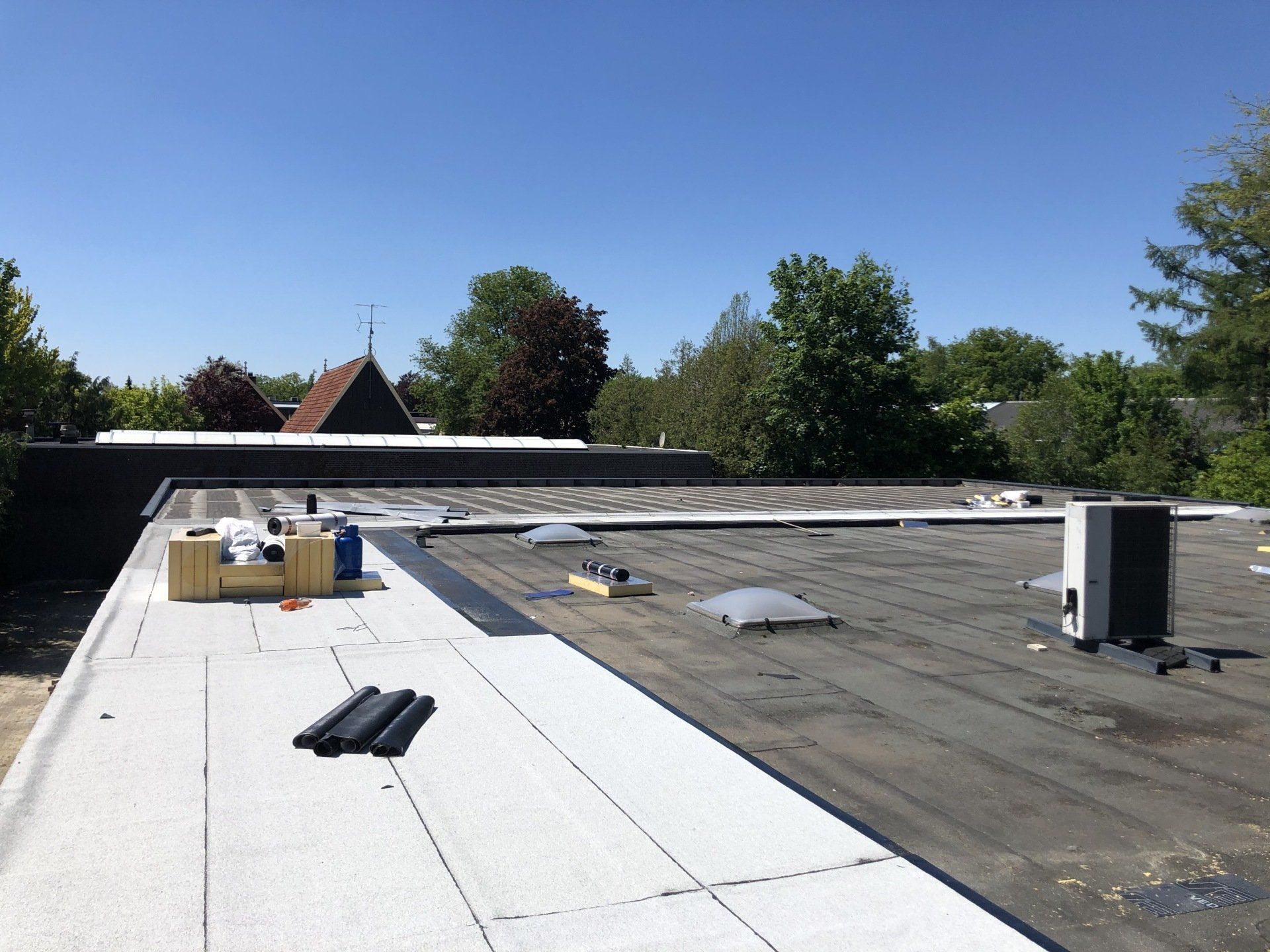 Een plat dak onder een helderblauwe hemel, waarop te zien is hoe een gedeelte van het nieuwe witte dakbedekkingsmateriaal wordt aangebracht nabij het oude bitumen.