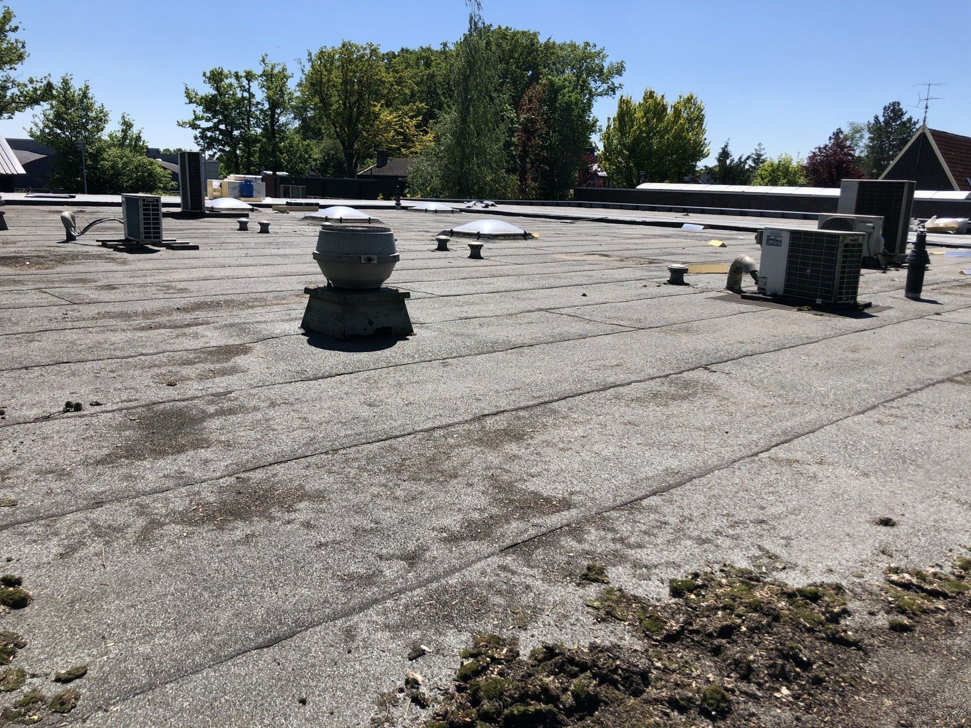 Een zicht op een plat, grijs bedrijfsdak met mechanische ventilatieopeningen, HVAC-units en hier en daar groene mosplekken.