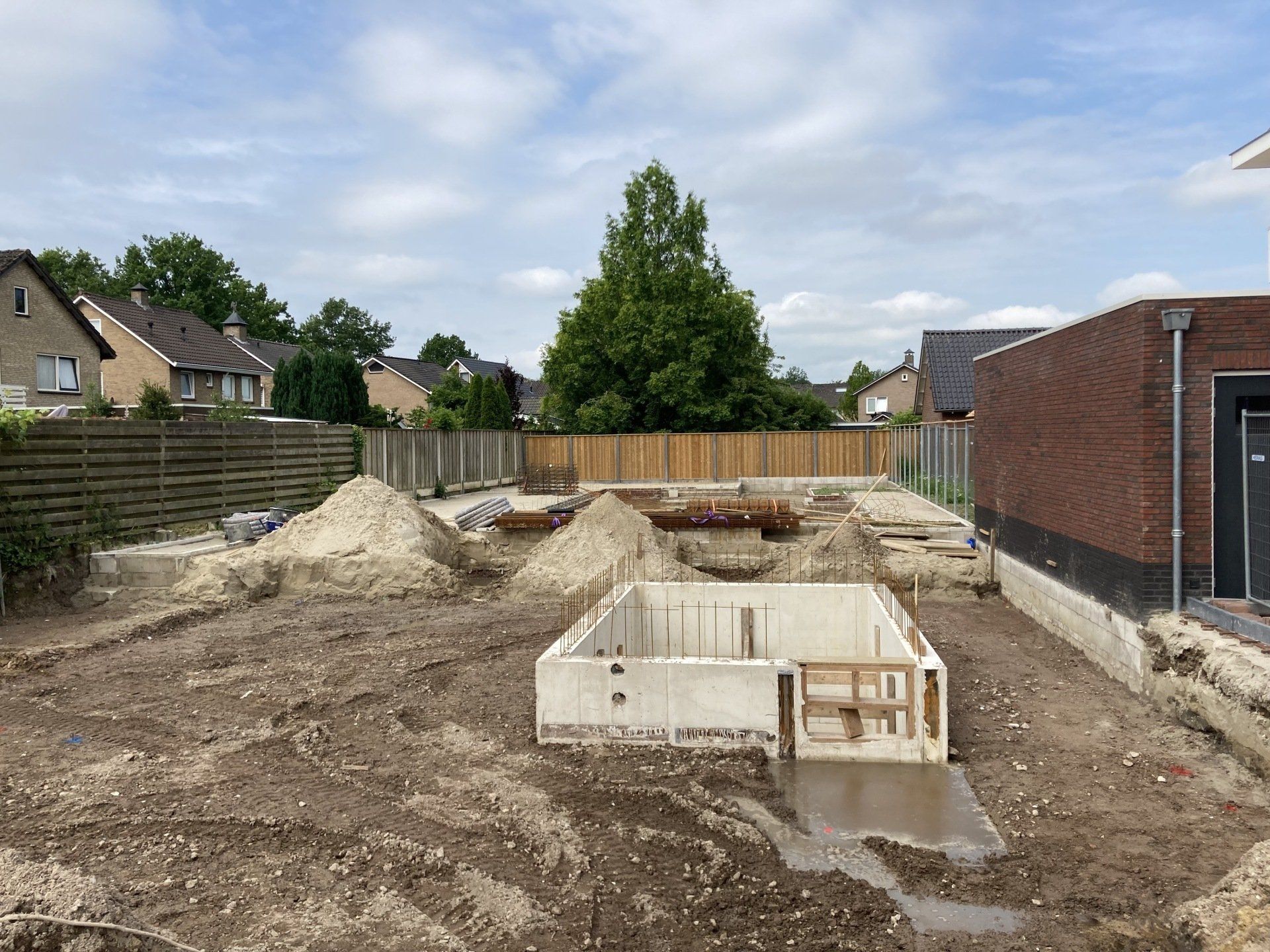 Bouwterrein met een rechthoekige betonnen fundering in een onverhard terrein naast een bakstenen gebouw.