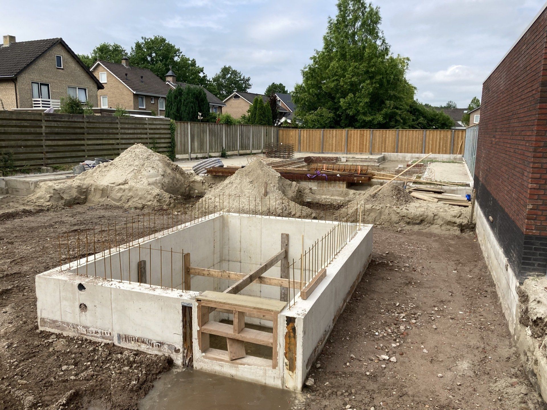 Een betonnen fundering voor een rechthoekige constructie in aanbouw in een achtertuin van een woonhuis, met hopen grond in de buurt.