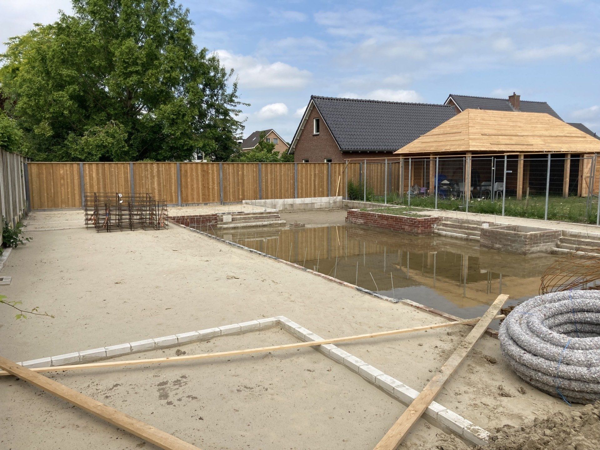 Een bouwterrein voor een woonhuis in de buitenlucht met een groot, halfvol zwembad, een zandbodem en een nieuw houten tuinhuisje.