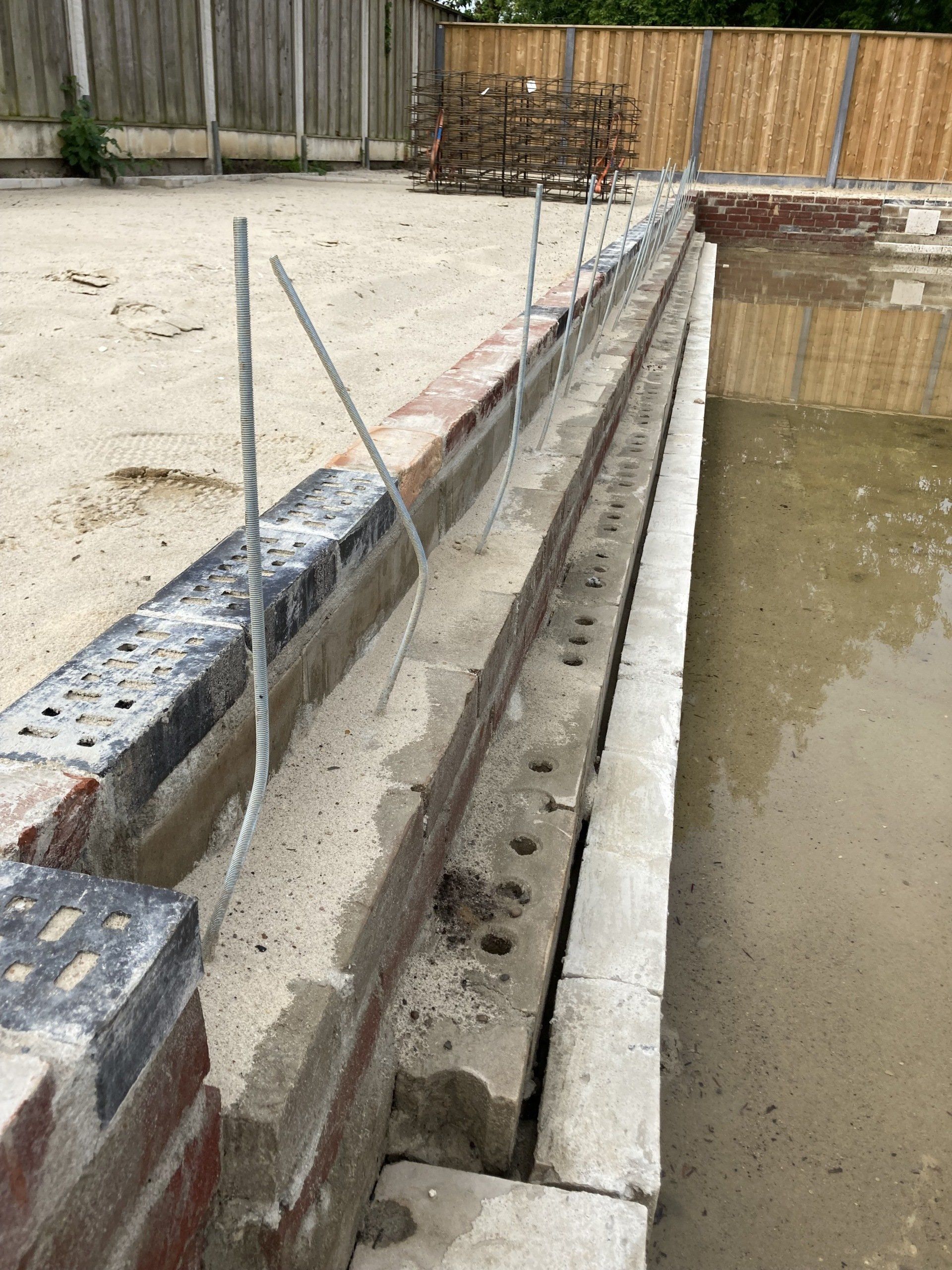 Een beeld van een bouwplaats met een half afgebouwde bakstenen muur en metalen steunbalken langs de rand van een met modder gevuld bassin.