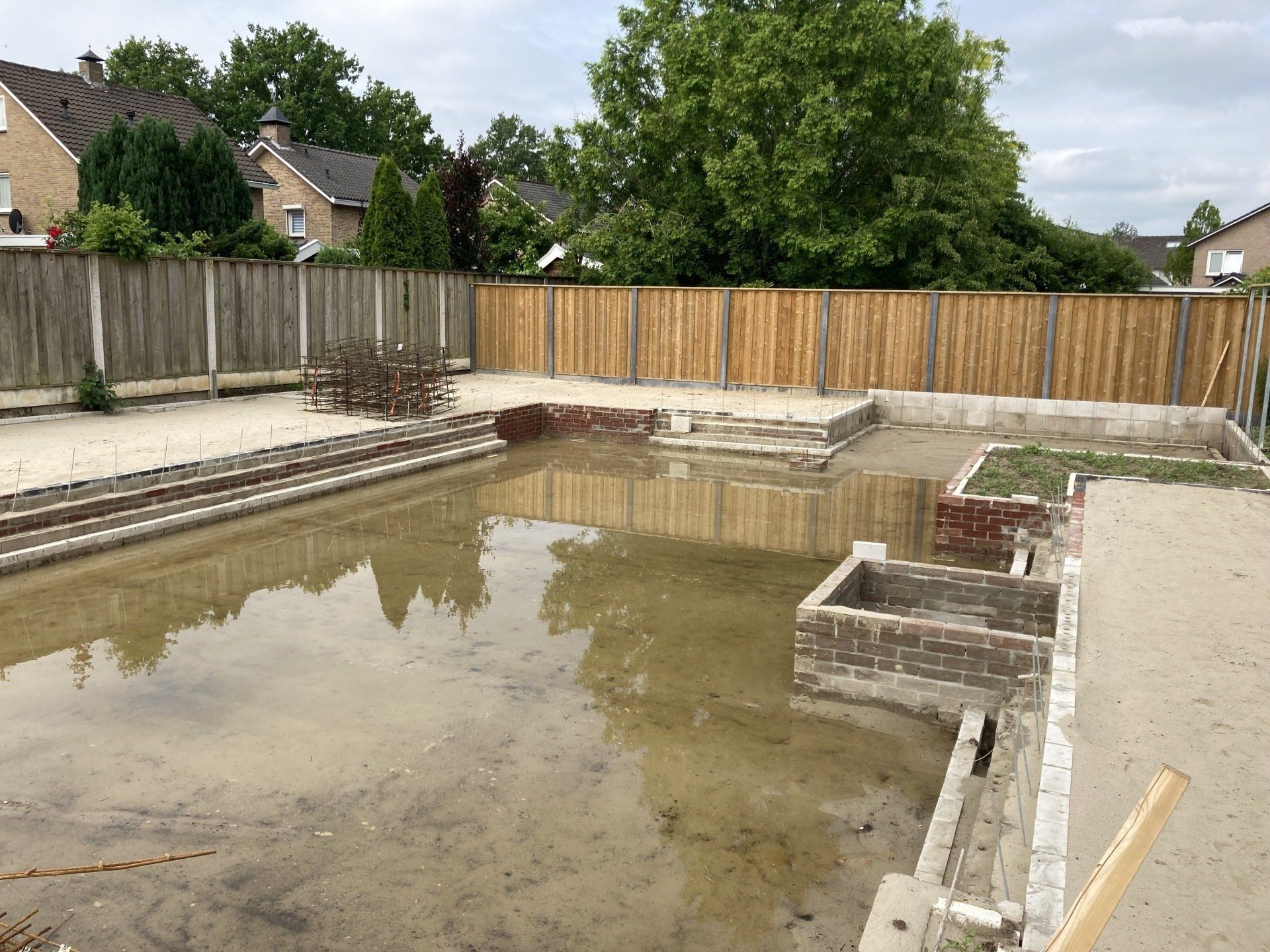 Een ondergelopen bouwterrein in een achtertuin met bakstenen fundering en houten schutting onder een bewolkte hemel.