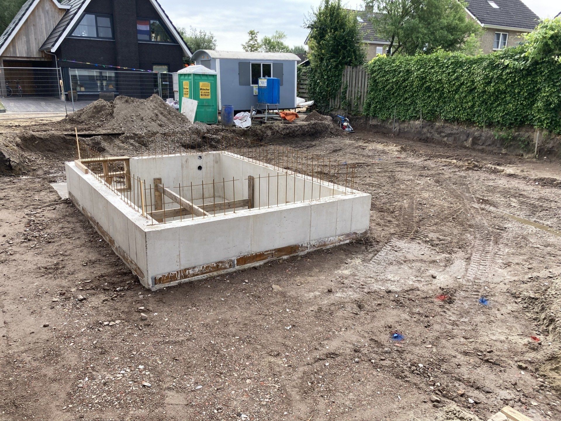 Op een stuk grond staat een betonnen fundering voor een klein gebouw, met op de achtergrond een huis en een klein schuurtje.