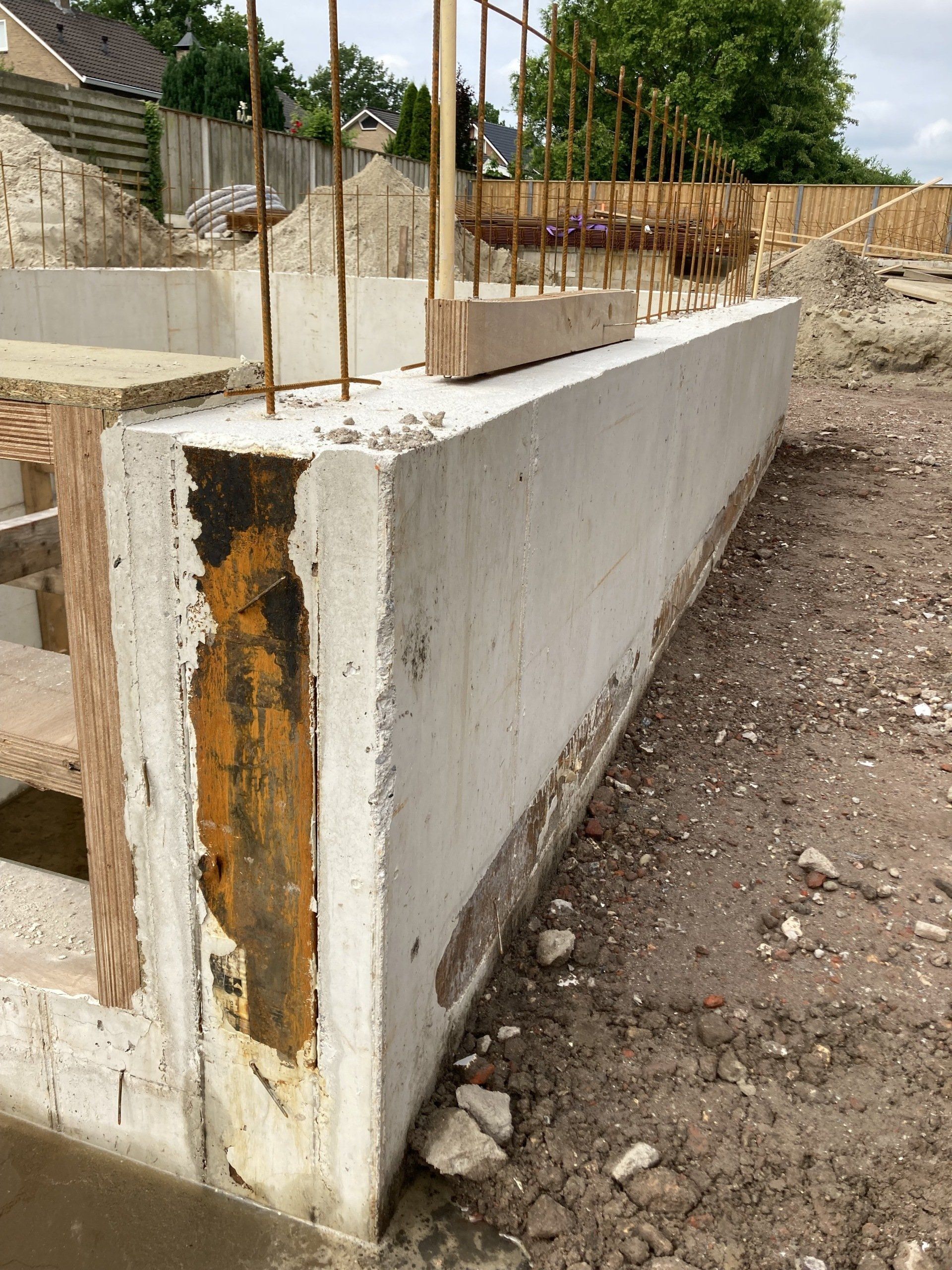 Een zijaanzicht van een pas gestorte betonnen funderingsmuur op een bouwterrein, met zichtbare verticale wapeningsstaven.