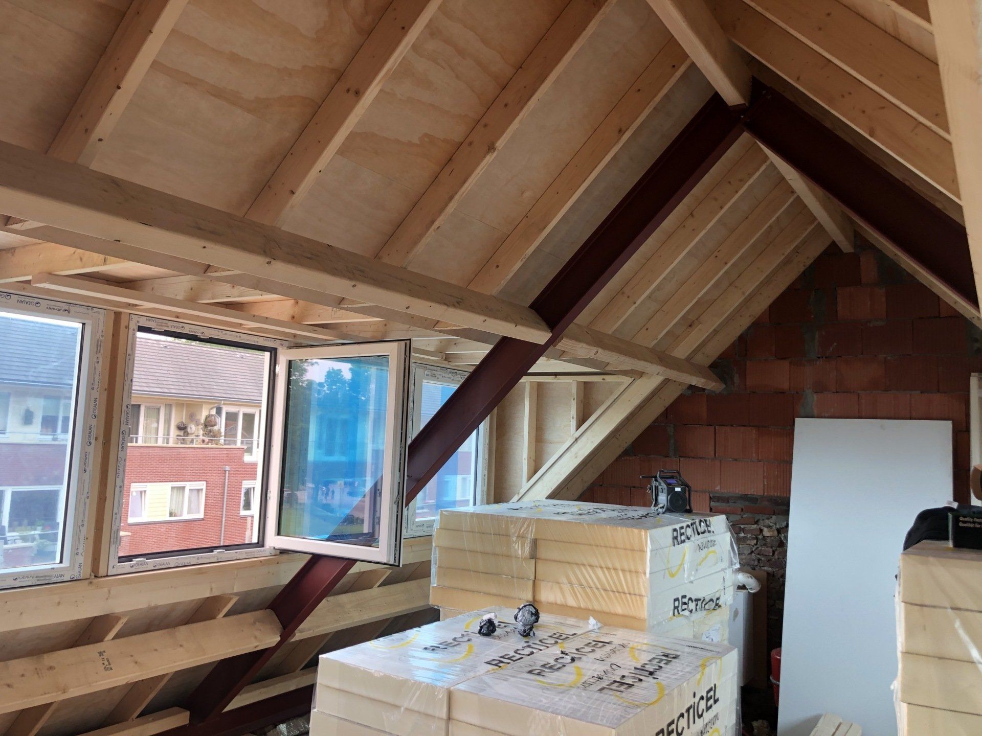 Een zolder in aanbouw met zichtbare houten balken, een raam, metalen steunbalken en stapels isolatiemateriaal.