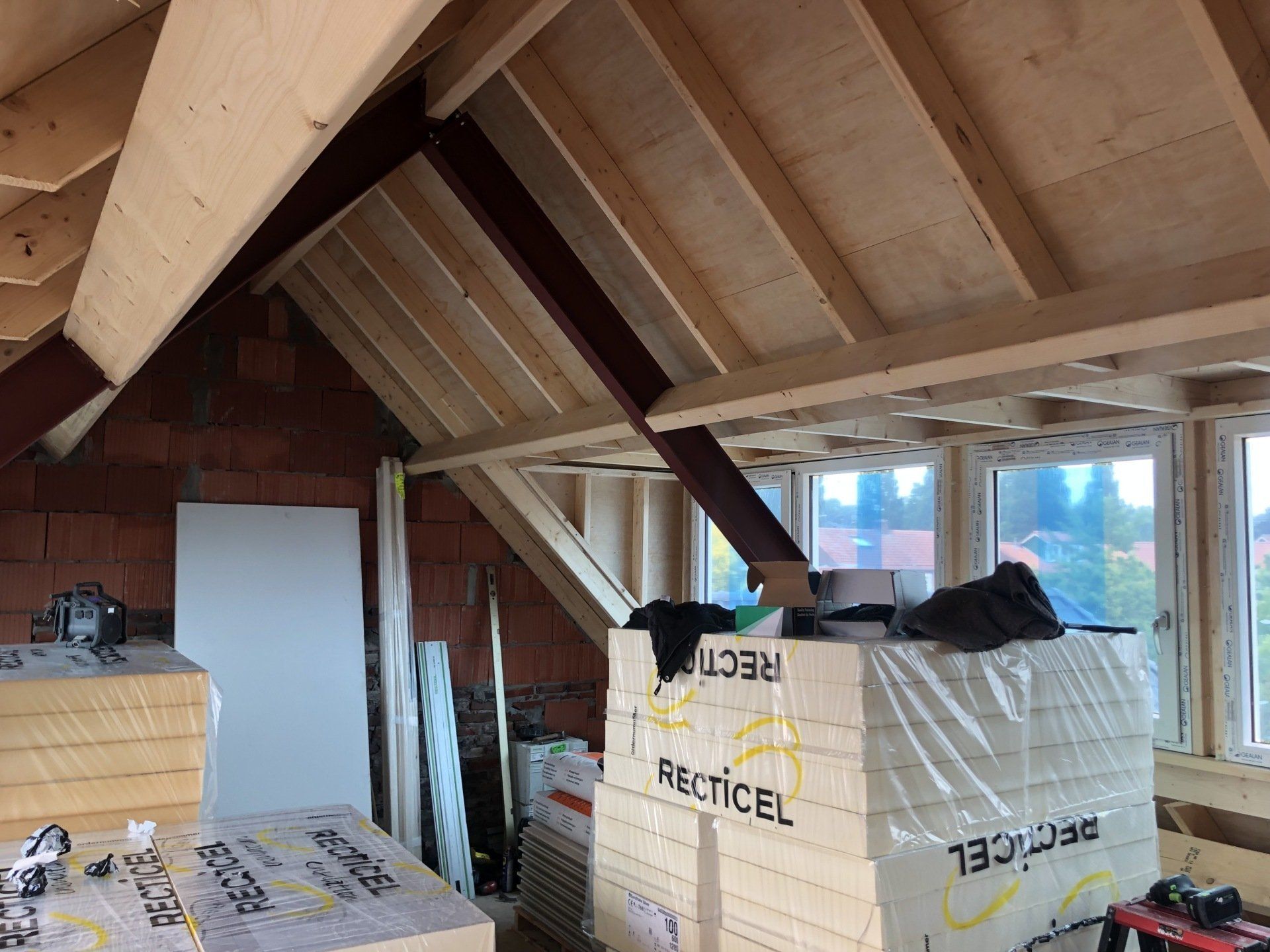 Een onafgewerkte zolderrenovatie met zichtbare houten dakbalken, bakstenen muren en stapels isolatiepanelen bij de ramen.