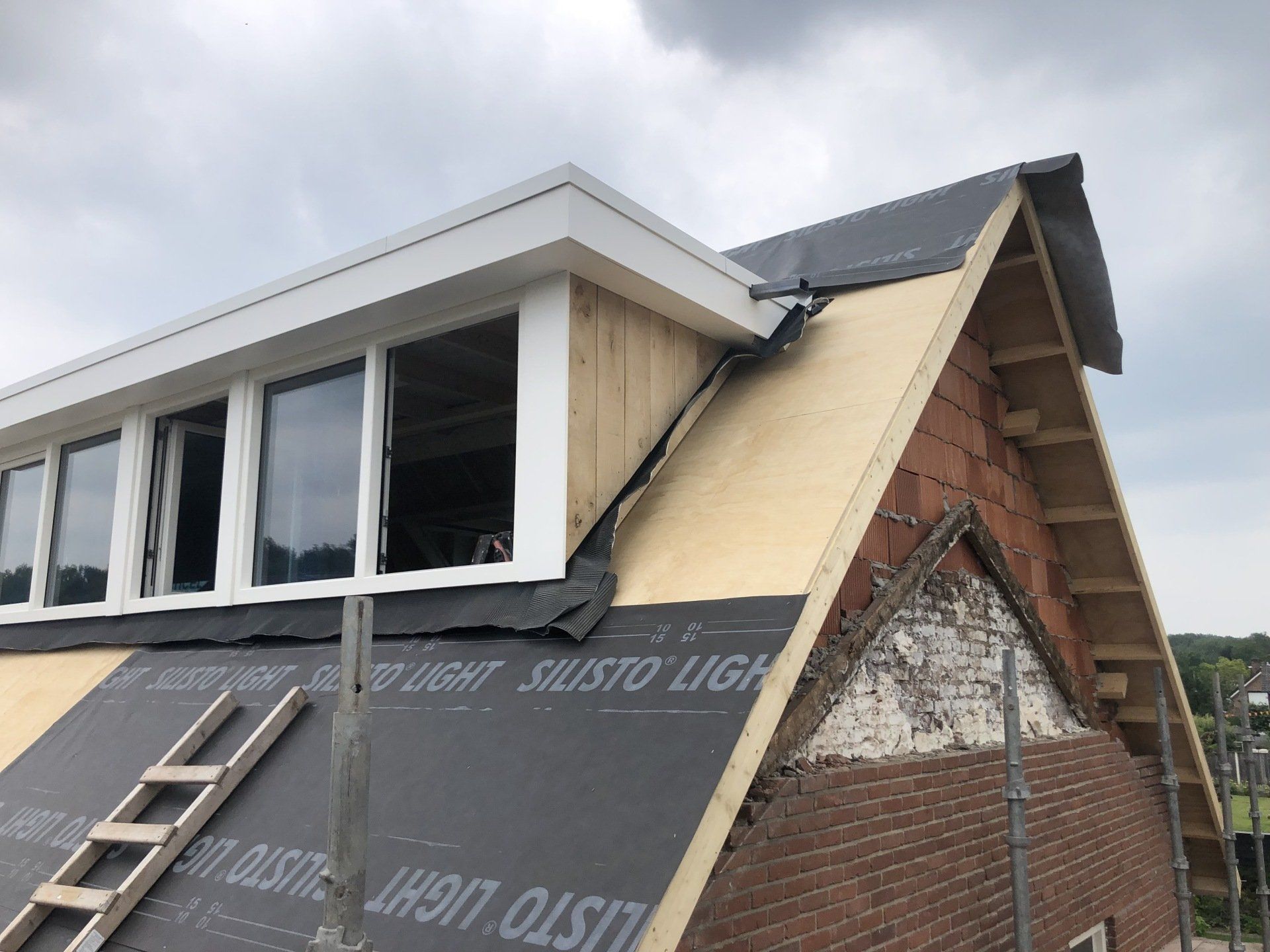 De constructie van een dakkapel op een hellend dak, met houten frame, donkere beschermlaag en buitenramen.
