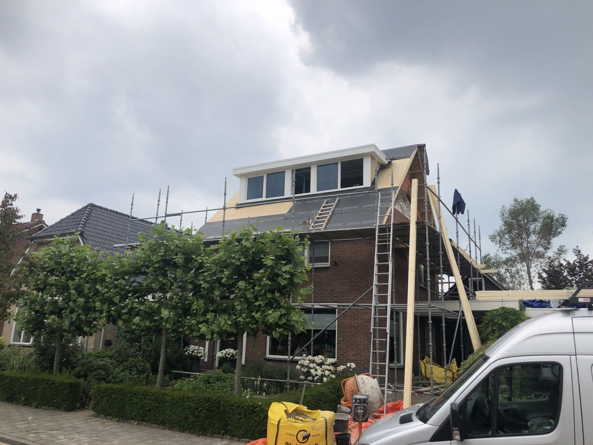 Een bakstenen huis in renovatie met een grote dakkapel, steigers en een ladder tegen het dak.
