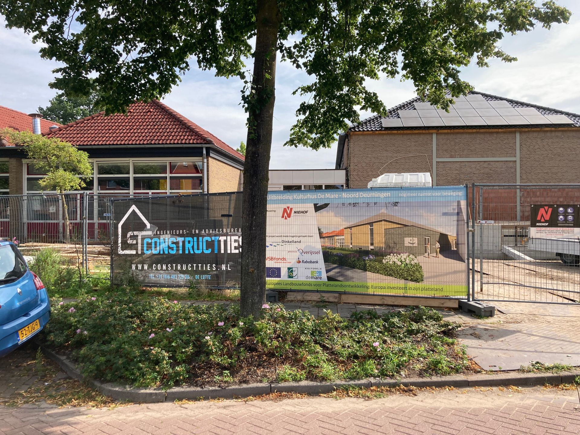 Een bouwterrein met een bord waarop bouwplannen te zien zijn, achter een hek van gaas, gezien vanaf de straat.
