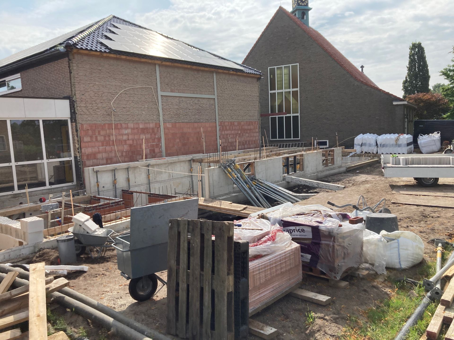 Bouwplaats met zichtbare betonnen funderingen, bouwmaterialen en steigers in de buurt van bakstenen gebouwen.
