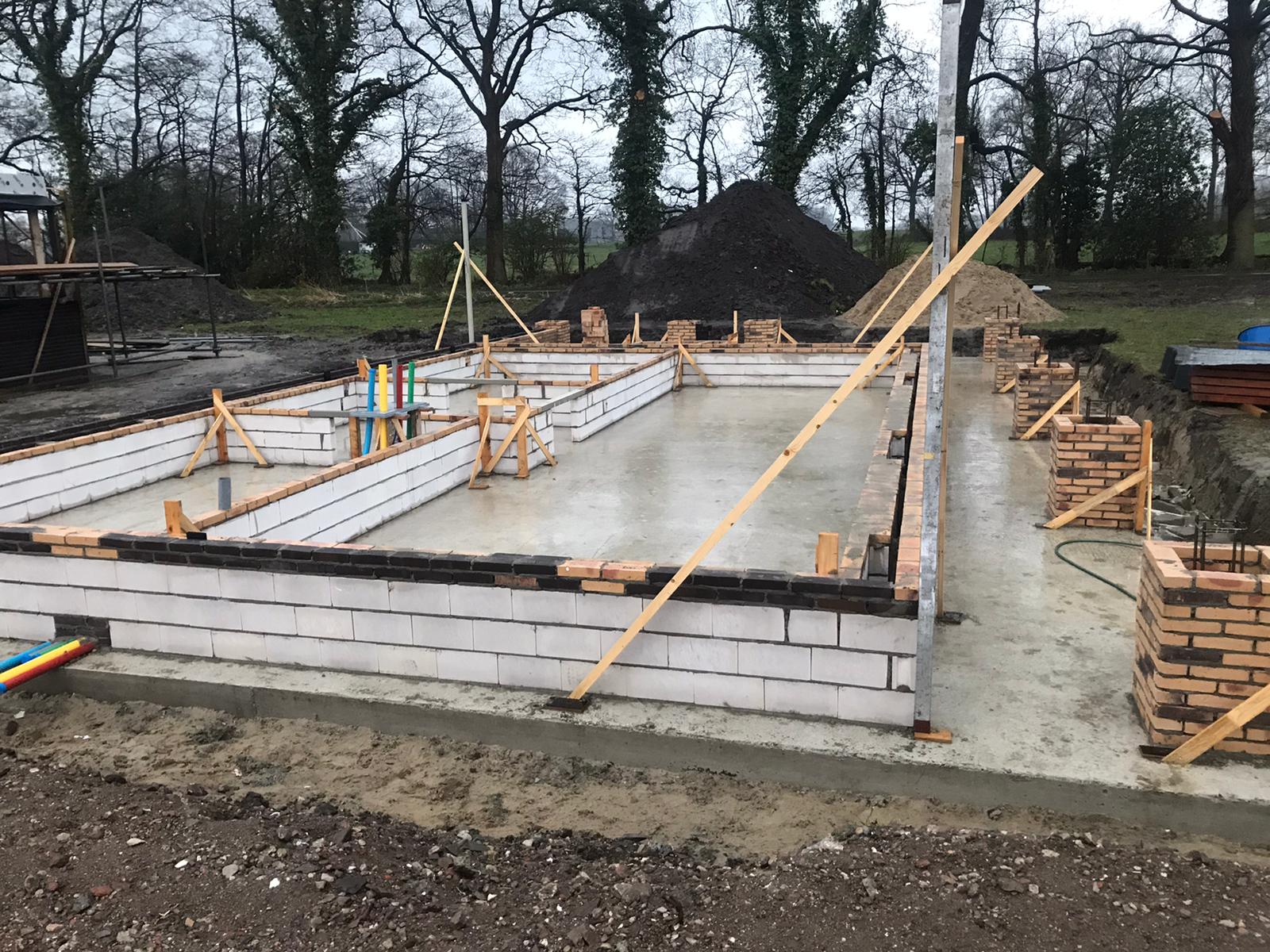 Een bouwplaats met funderingen van betonblokken en een gestorte betonplaat op een landelijk perceel met bomen op de achtergrond.