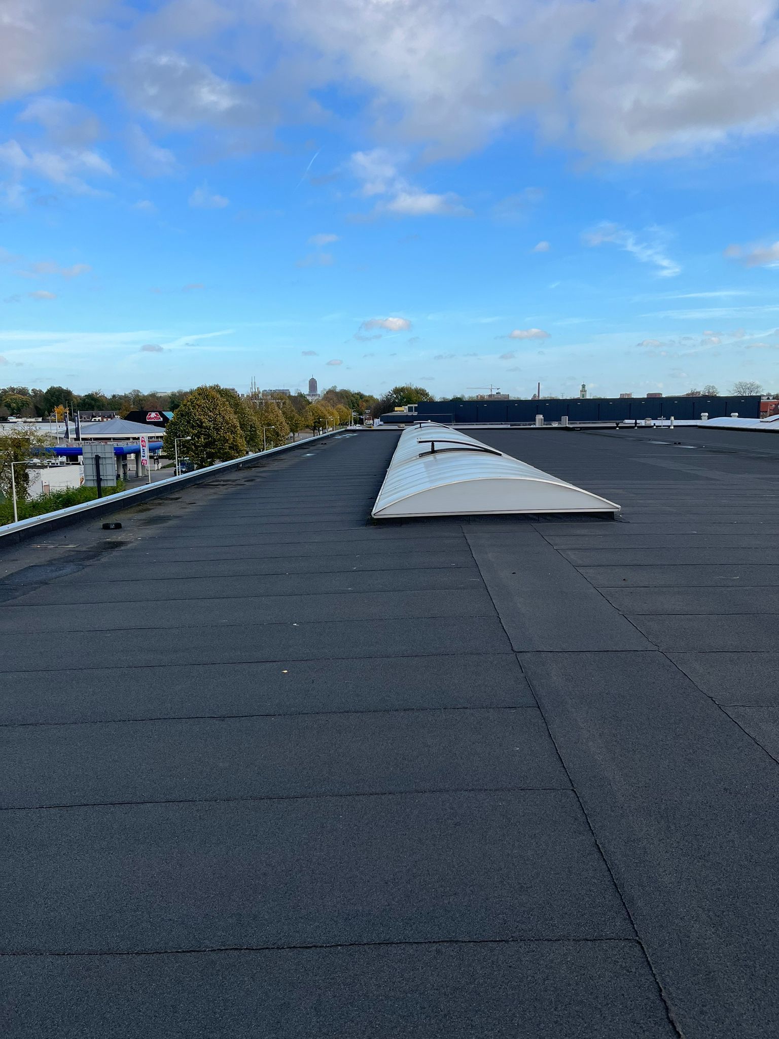Een plat, donker grinddak onder een blauwe, bewolkte hemel, met een lang, gebogen dakraam in het midden.