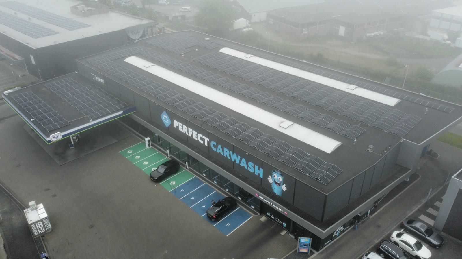Een luchtfoto van een zwarte Perfect Carwash-vestiging met zonnepanelen op het dak en een parkeerplaats met laadpalen.
