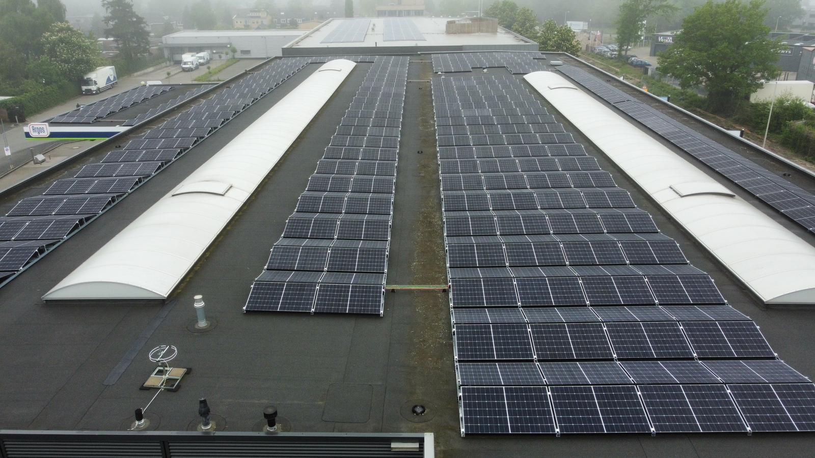 Een luchtfoto van een plat dak met uitgebreide rijen donkere zonnepanelen, gescheiden door witte dakramen.