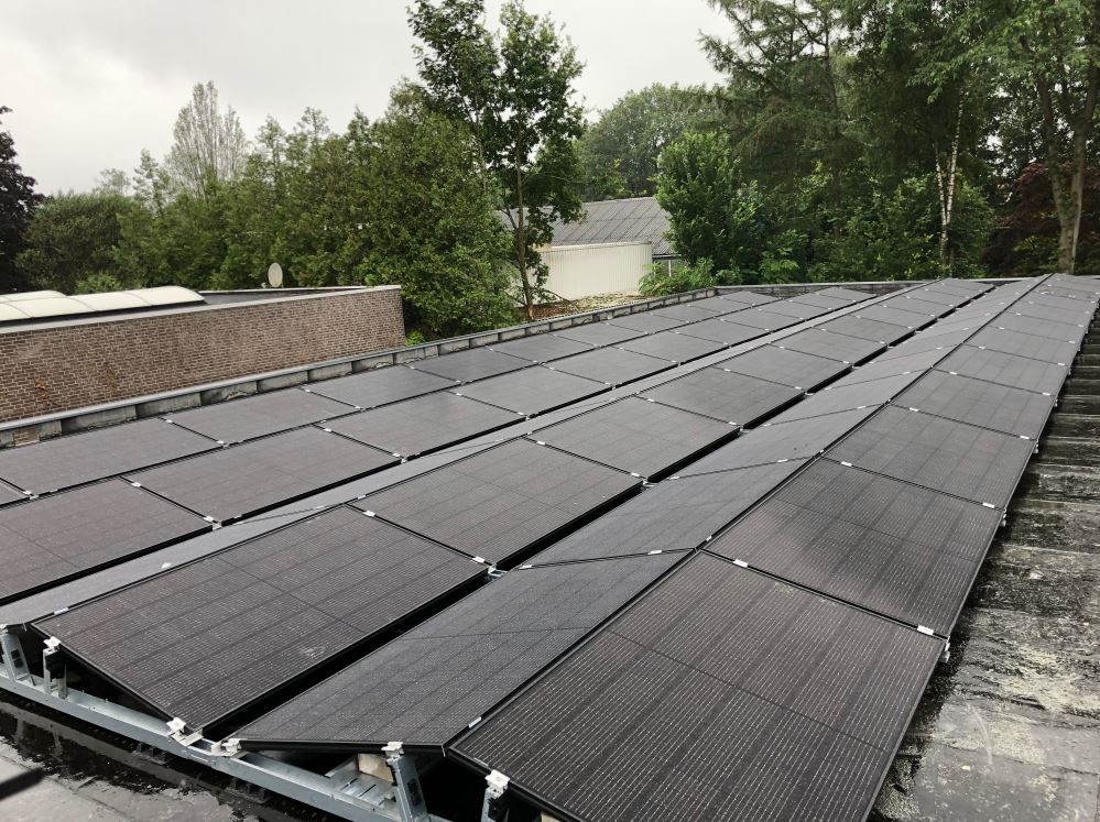 Rijen donkere zonnepanelen geïnstalleerd op een plat dak tegen een achtergrond van groene bomen en een bewolkte hemel.