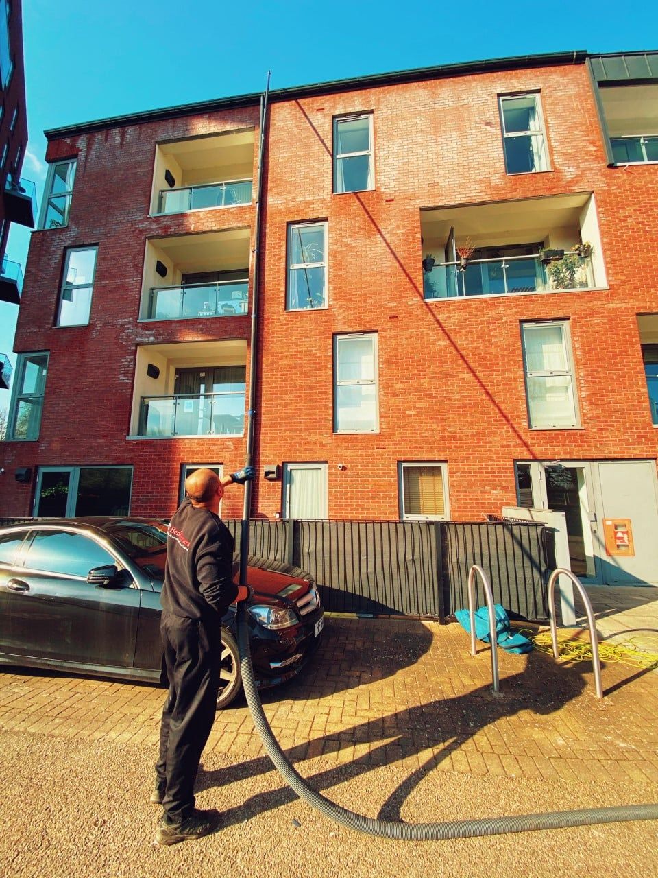 Man cleaning windows on a brick apartment building with a hose, parked cars nearby.