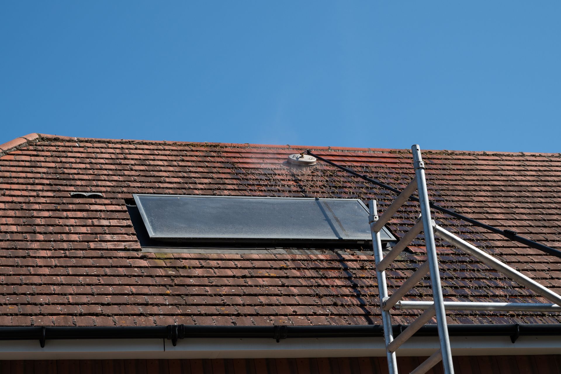 Roof being cleaned with power washer, brown tiles, blue sky, and scaffolding present.