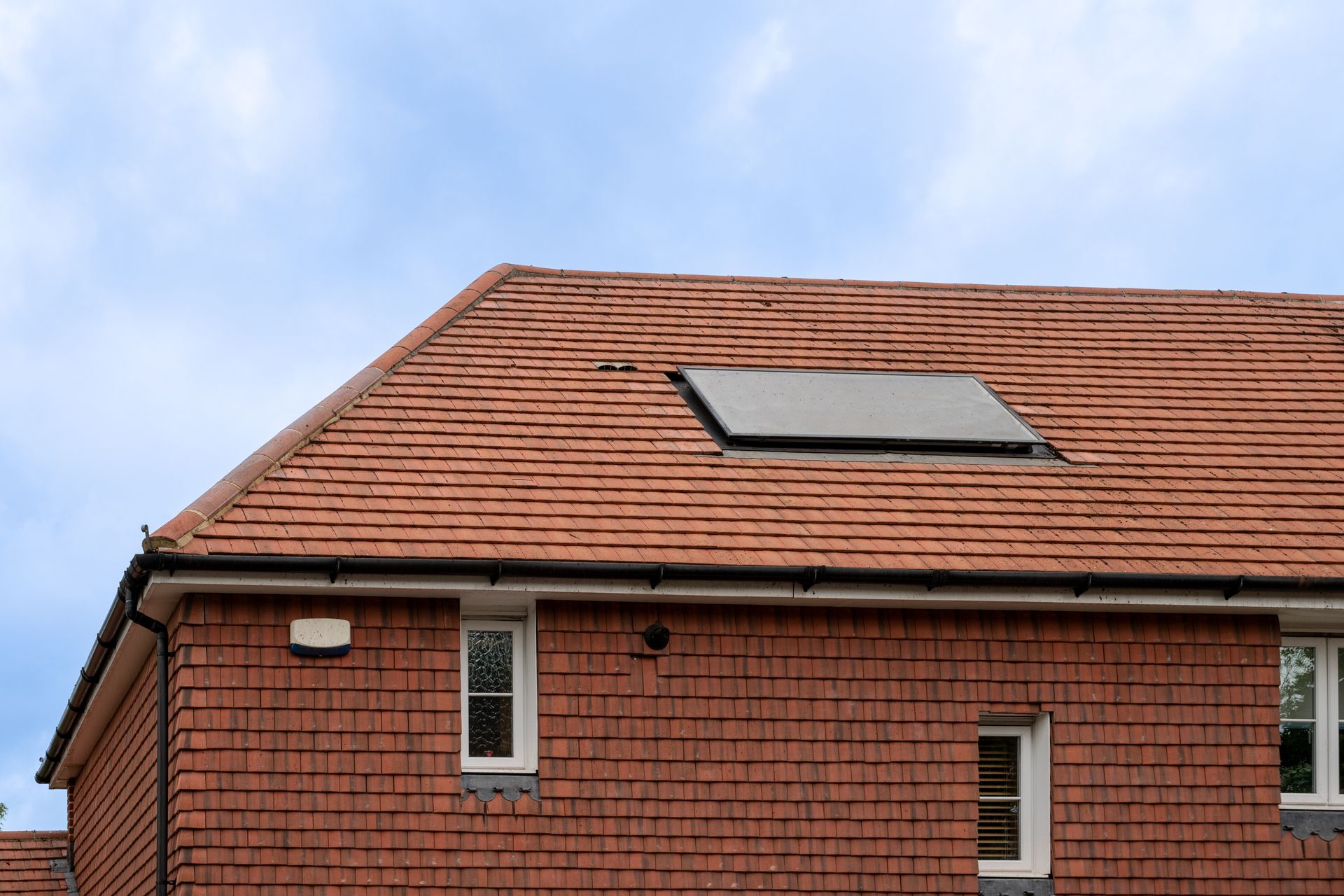 Brick building with red tiled roof, rectangular skylight, and small windows against a cloudy blue sky.