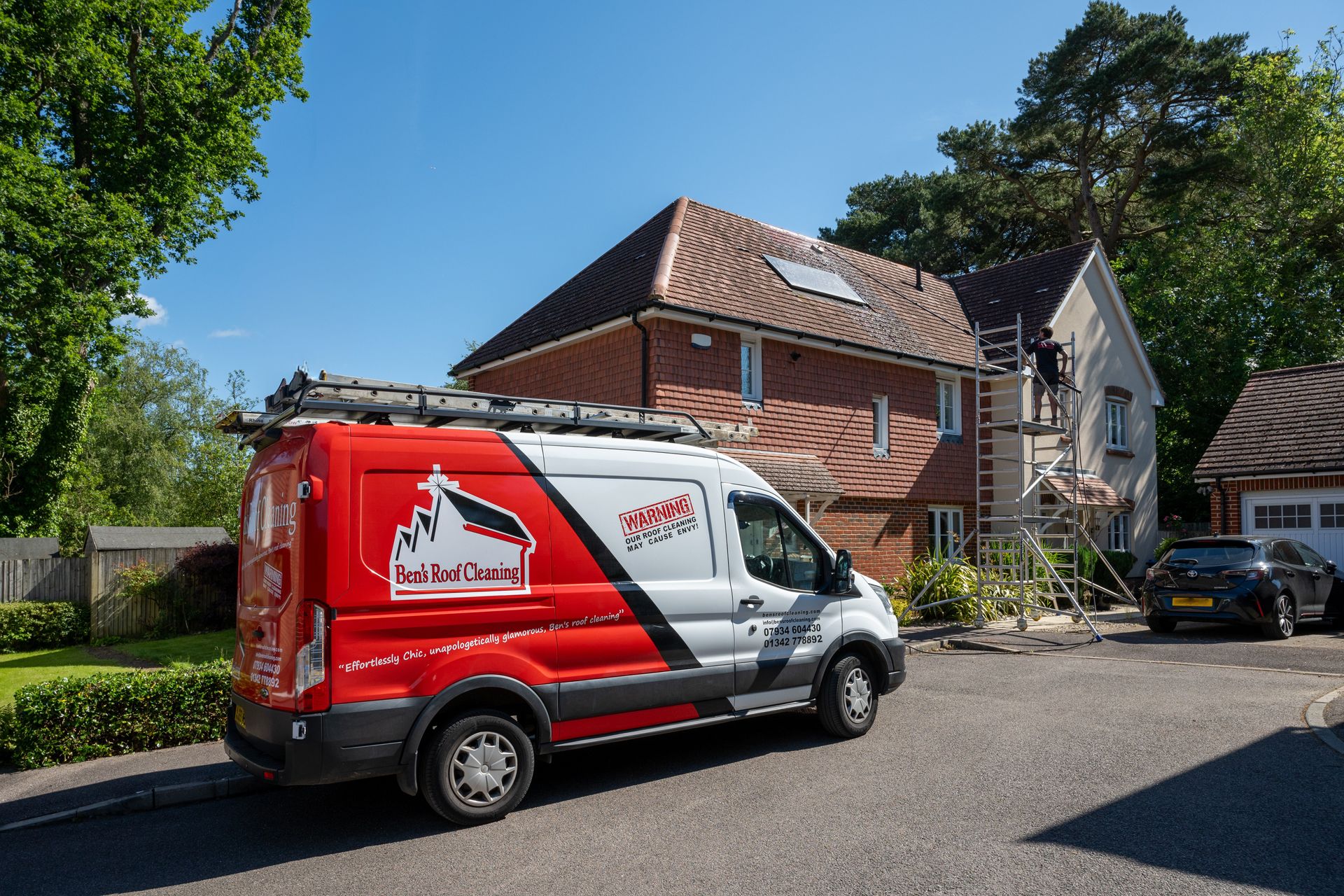 Red and white service van parked in front of a house, with scaffolding and a ladder on the roof.