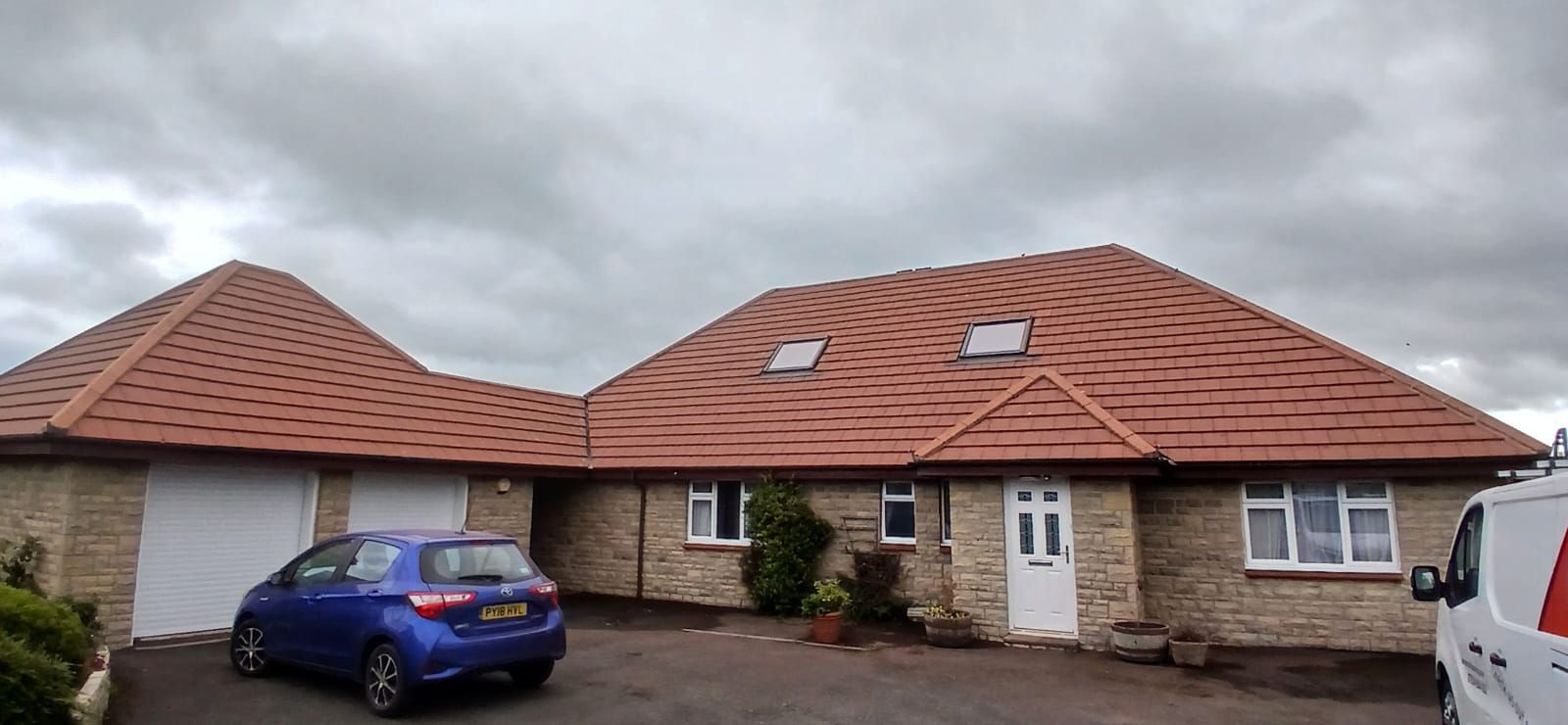 A light stone house with a red tiled roof, blue car in the driveway, and a cloudy sky.