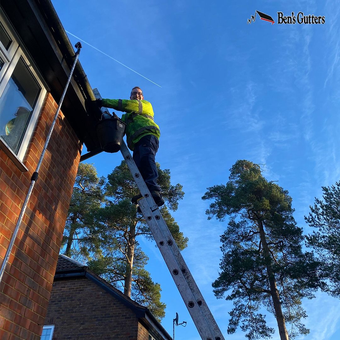 Person on ladder cleaning gutters; bright clothing against blue sky. Brick house, trees.