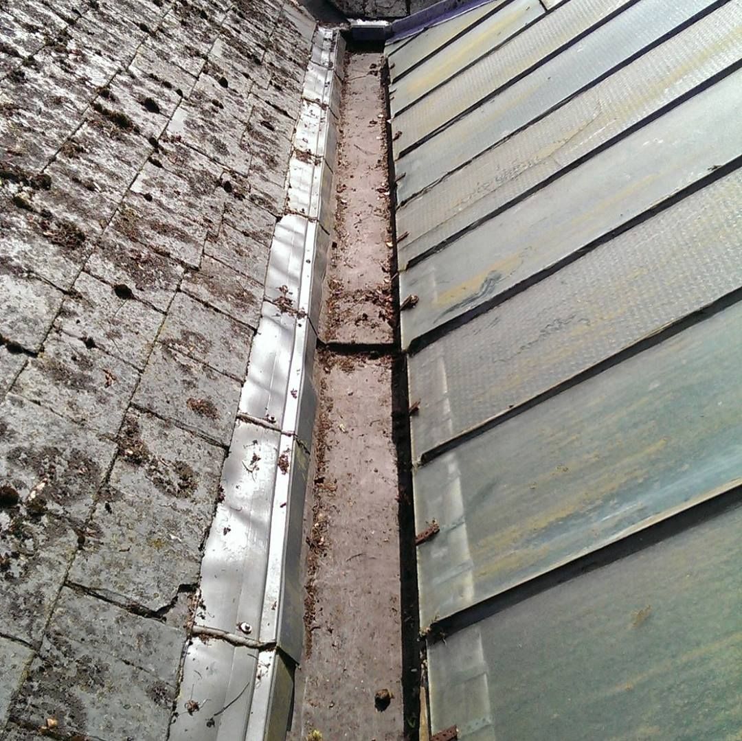 Gutter between a brick wall and a roof with green-gray metal tiles; debris covers the gutter.