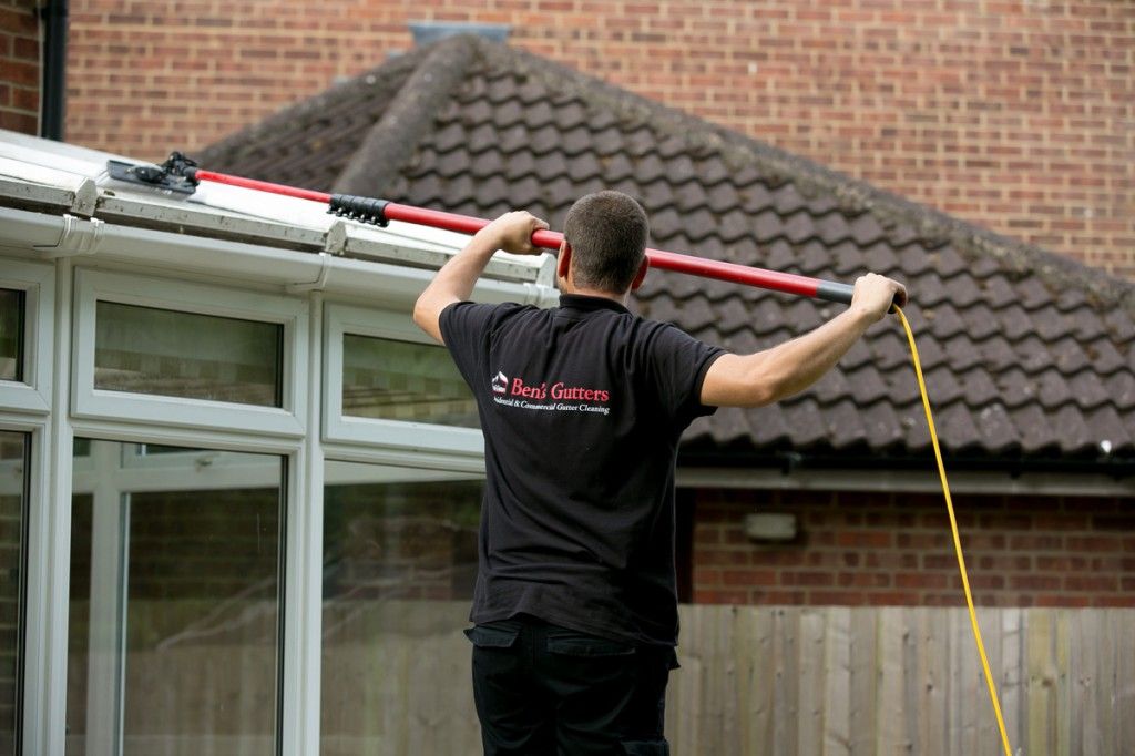 Man cleaning gutters with a long-handled tool on a house with white windows and a brown roof.