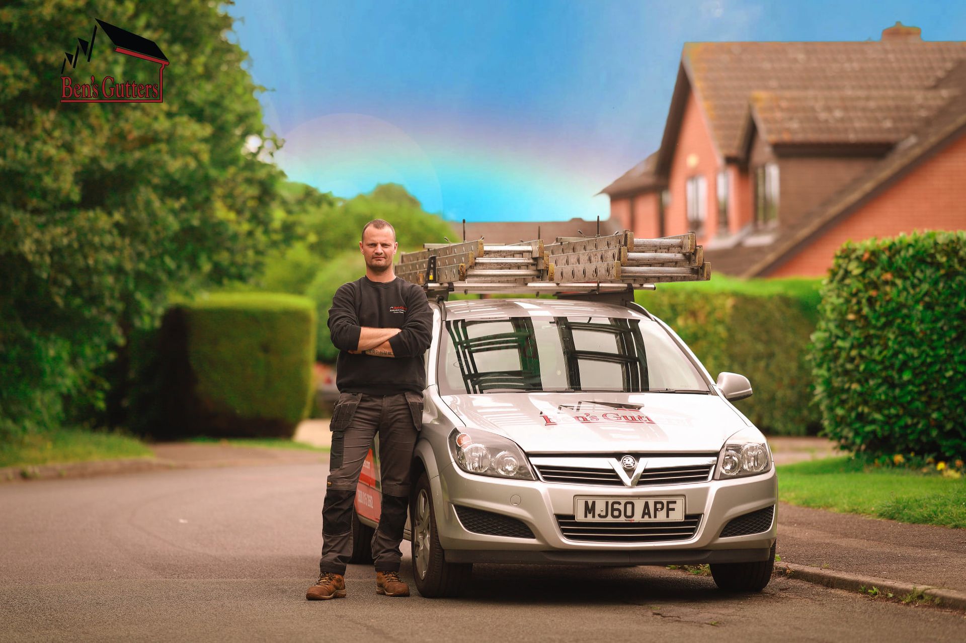 Man stands by silver van with ladder on roof; rainbow in sky.