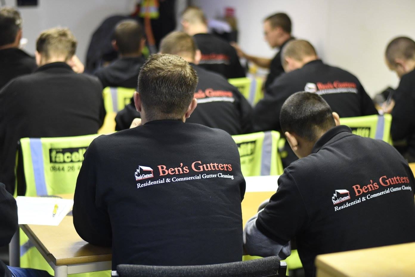 People in black sweatshirts with logos seated at tables in a room, presumably a training session.