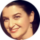 Smiling person in a circular portrait with dark hair and dangling earrings, softly lit against a light background