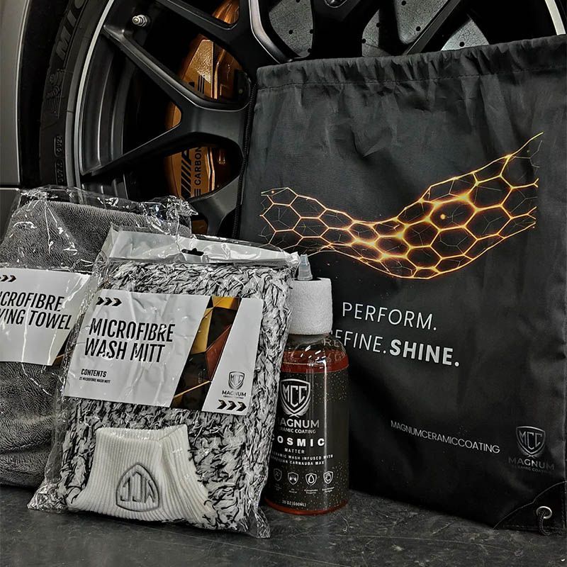 Car care kit with microfiber wash mitt and towel, wheel brush, and black branded bag beside a car wheel.